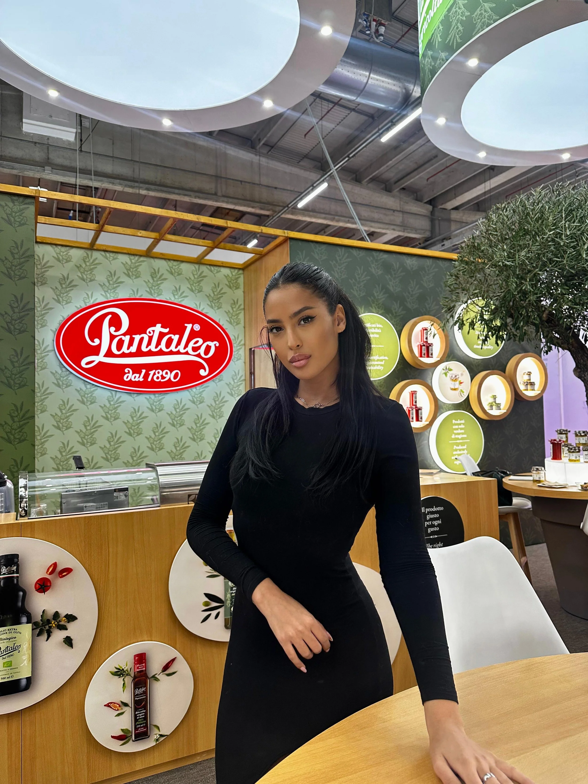Una donna con capelli neri vestita di nero davanti a uno stand con il logo 'Pantaleo dal 1890', decorato con prodotti e foglie di ulivo, all'interno di un ambiente ben illuminato.