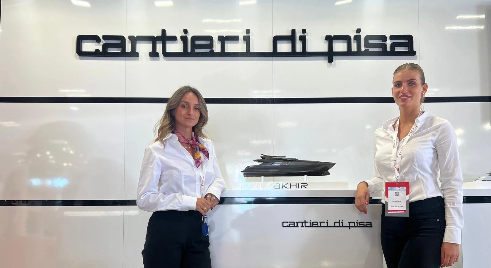 Due donne in abiti formali davanti a una reception con il logo 'cantieri di pisa' sul muro alle spalle.