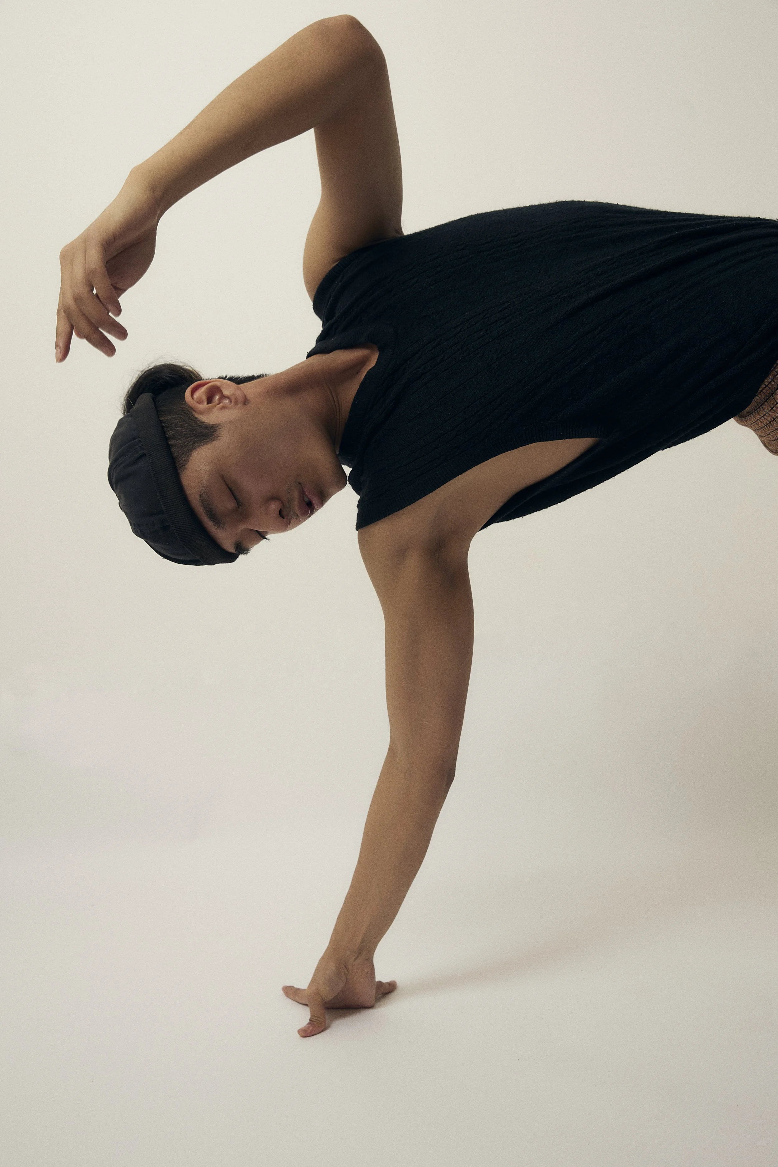 A danser perfomance, balancing on one hand with one arm bent and their head bowed, dressed in a black sleeveless top, with a plain white background.