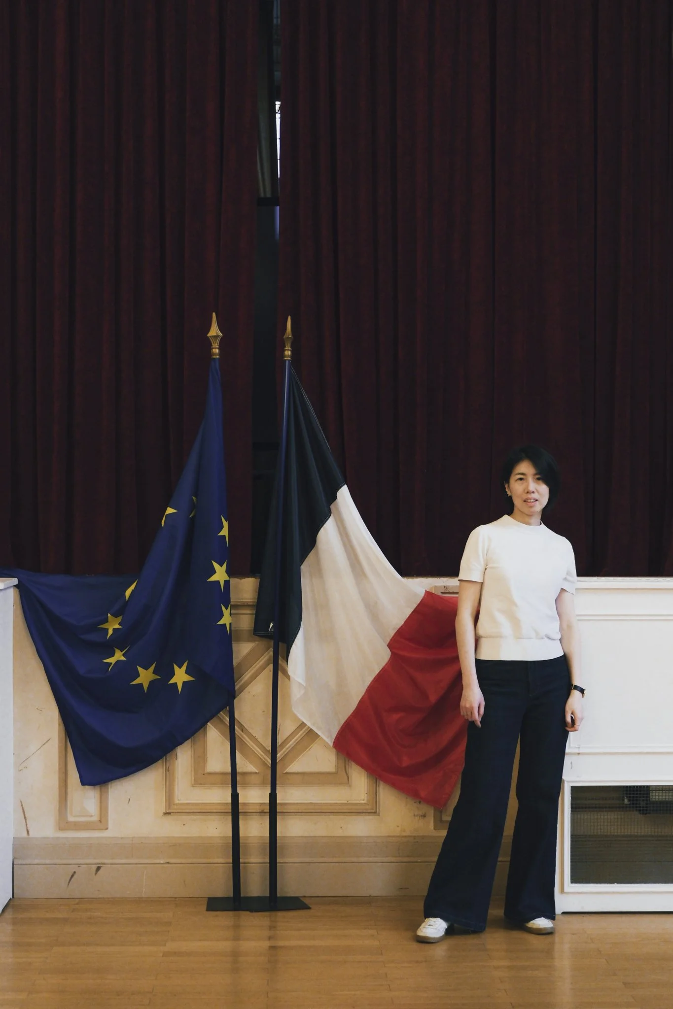 A woman standing next to two flags, the European Union flag and the French flag, in front of a dark red curtain. It's the Ceremony of French Nationality.