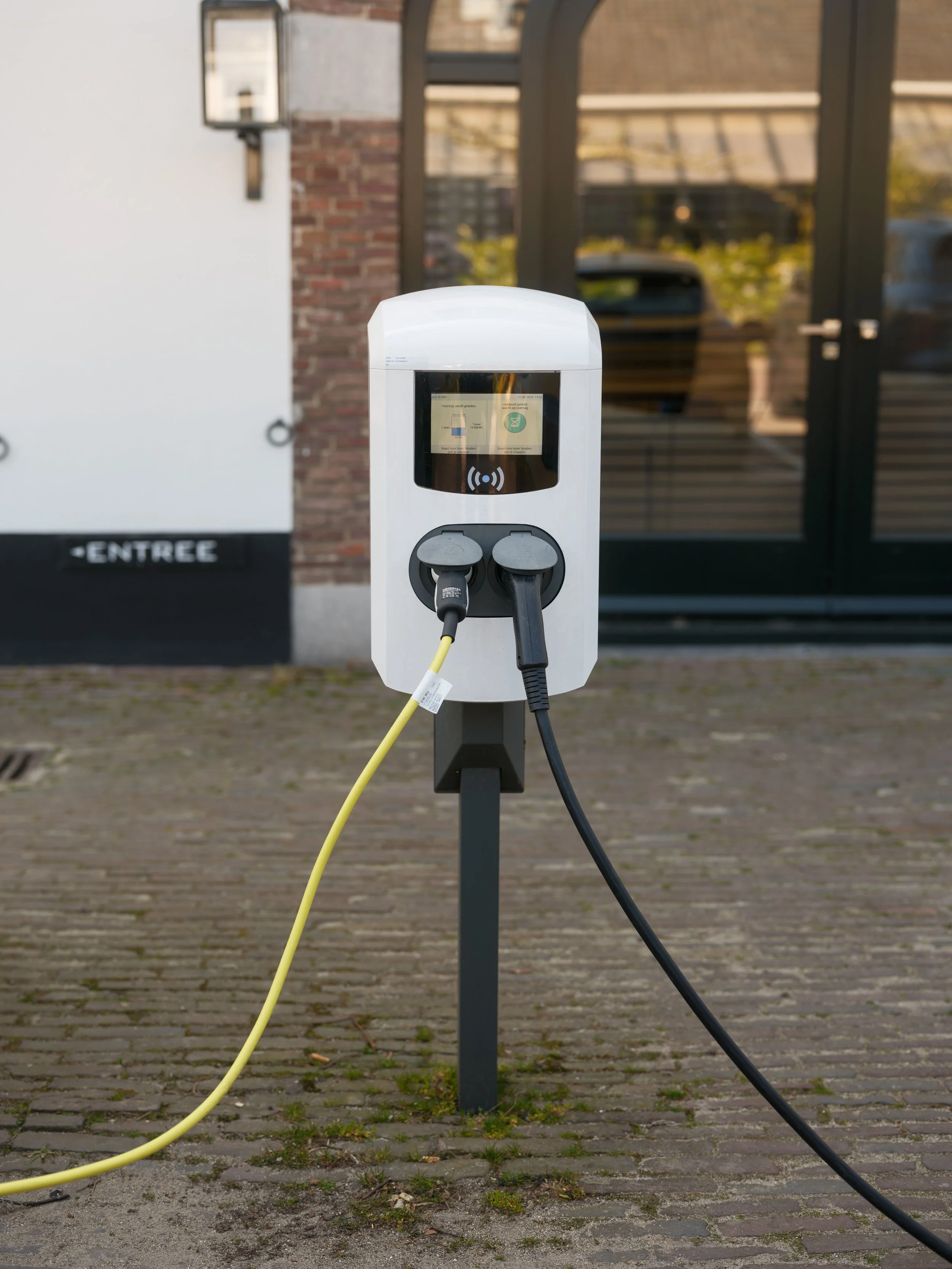 Electric vehicle charging station with two charging cables, set outdoors on a paved area.