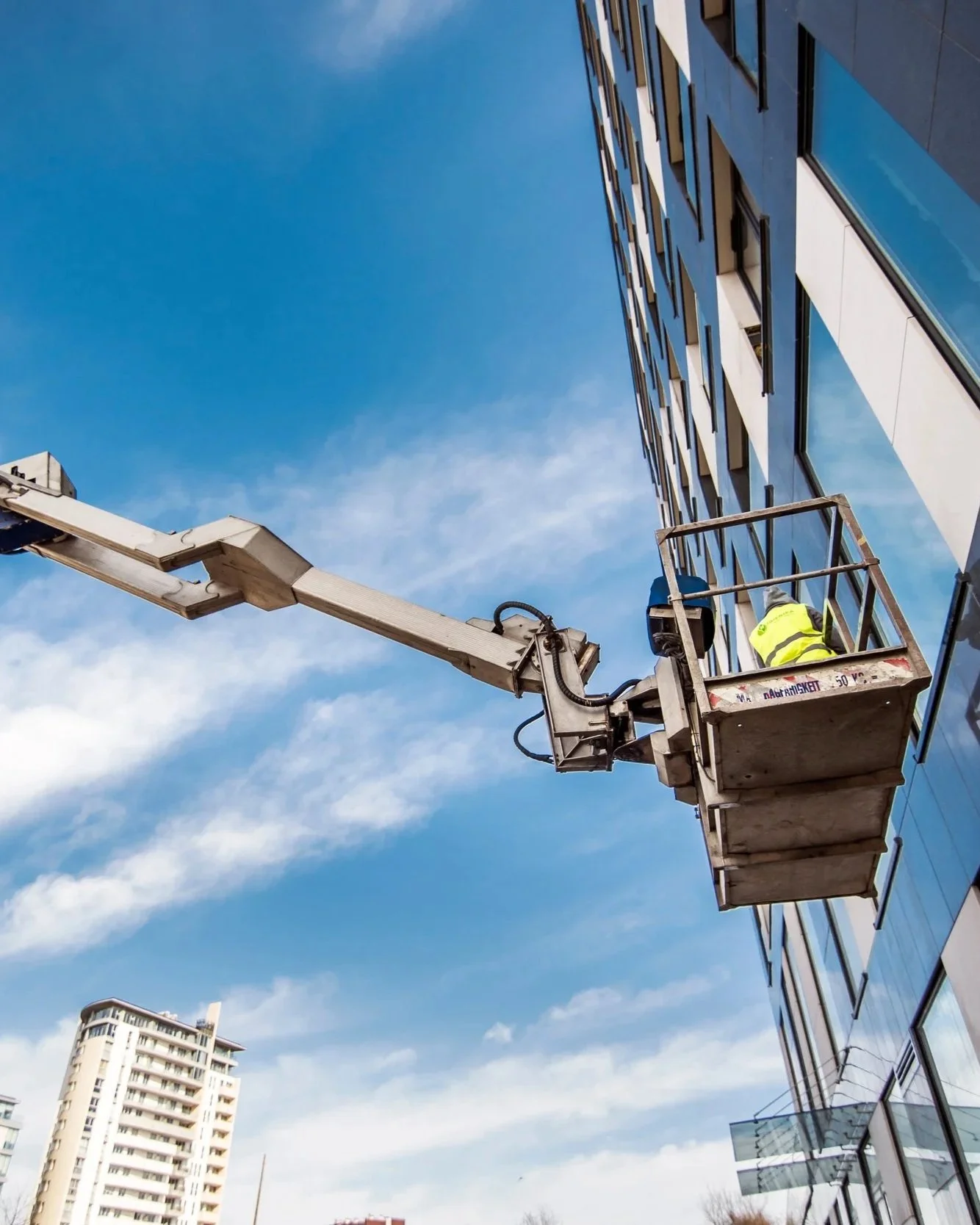 A worker in a yellow safety vest is on a cherry picker working on the exterior of a modern glass building. The cherry picker extends from the ground to the building's upper floors, with a clear blue sky in the background.