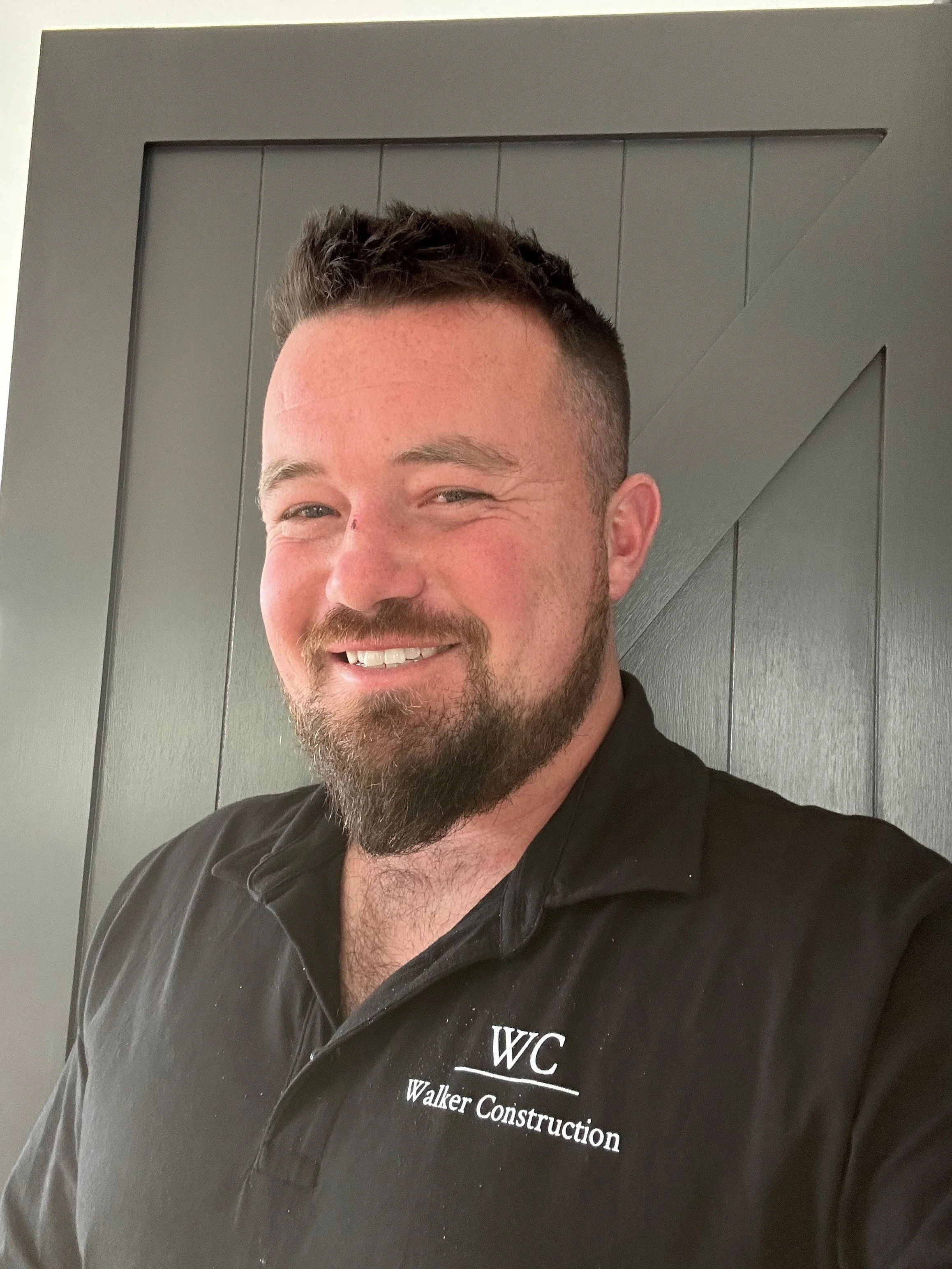 A man smiling, with short dark hair and a beard, wearing a black polo shirt with white text that reads 'WC Walker Construction', standing against a gray paneled wall inside a building.