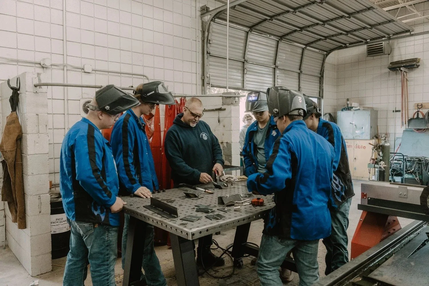 A group of students wearing blue work jackets and welding helmets around a worktable, with an instructor demonstrating welding techniques in a workshop.