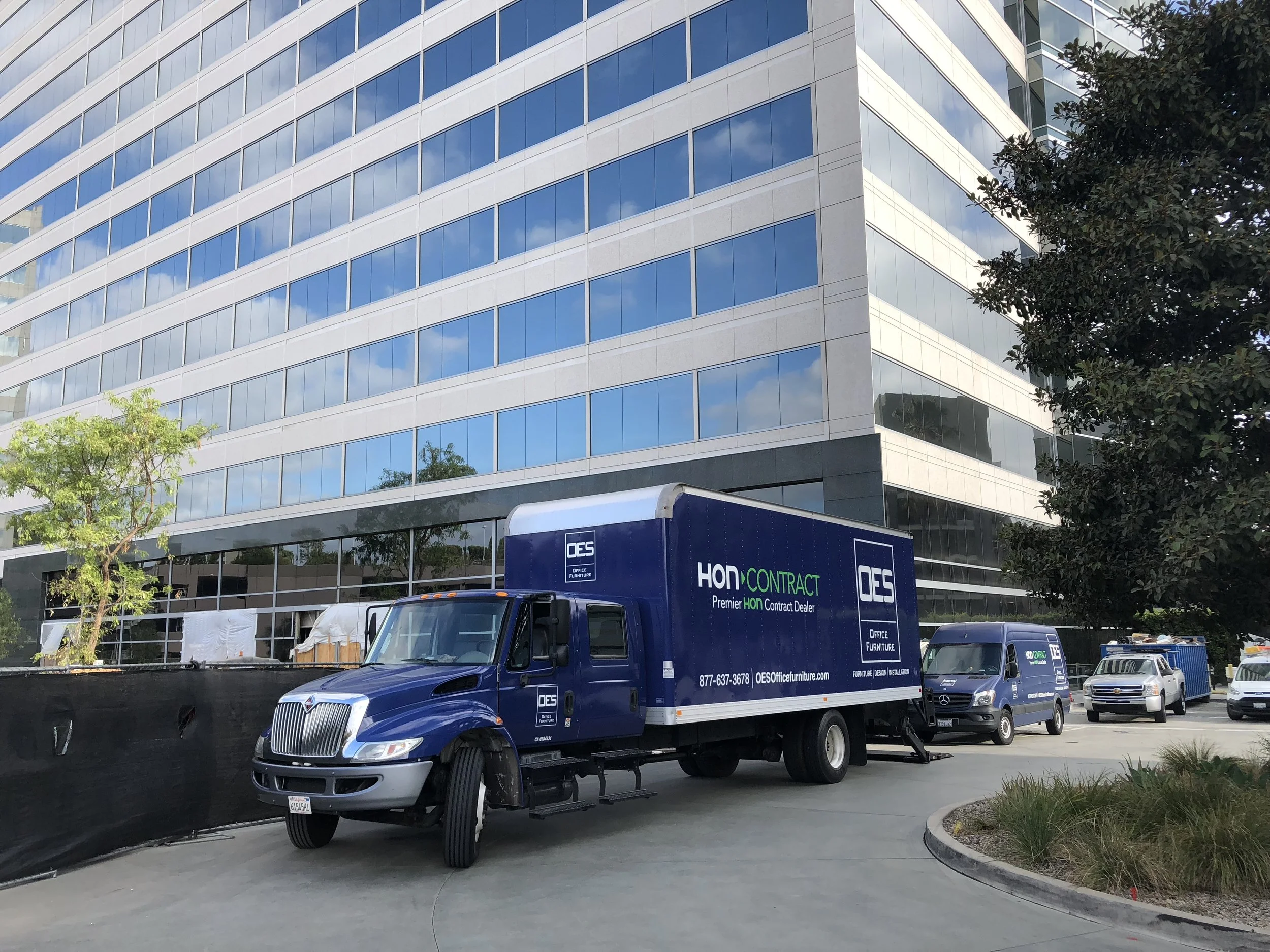 Blue Office Furniture delivery truck parked near a modern glass office building with trees and other vehicles in the background.