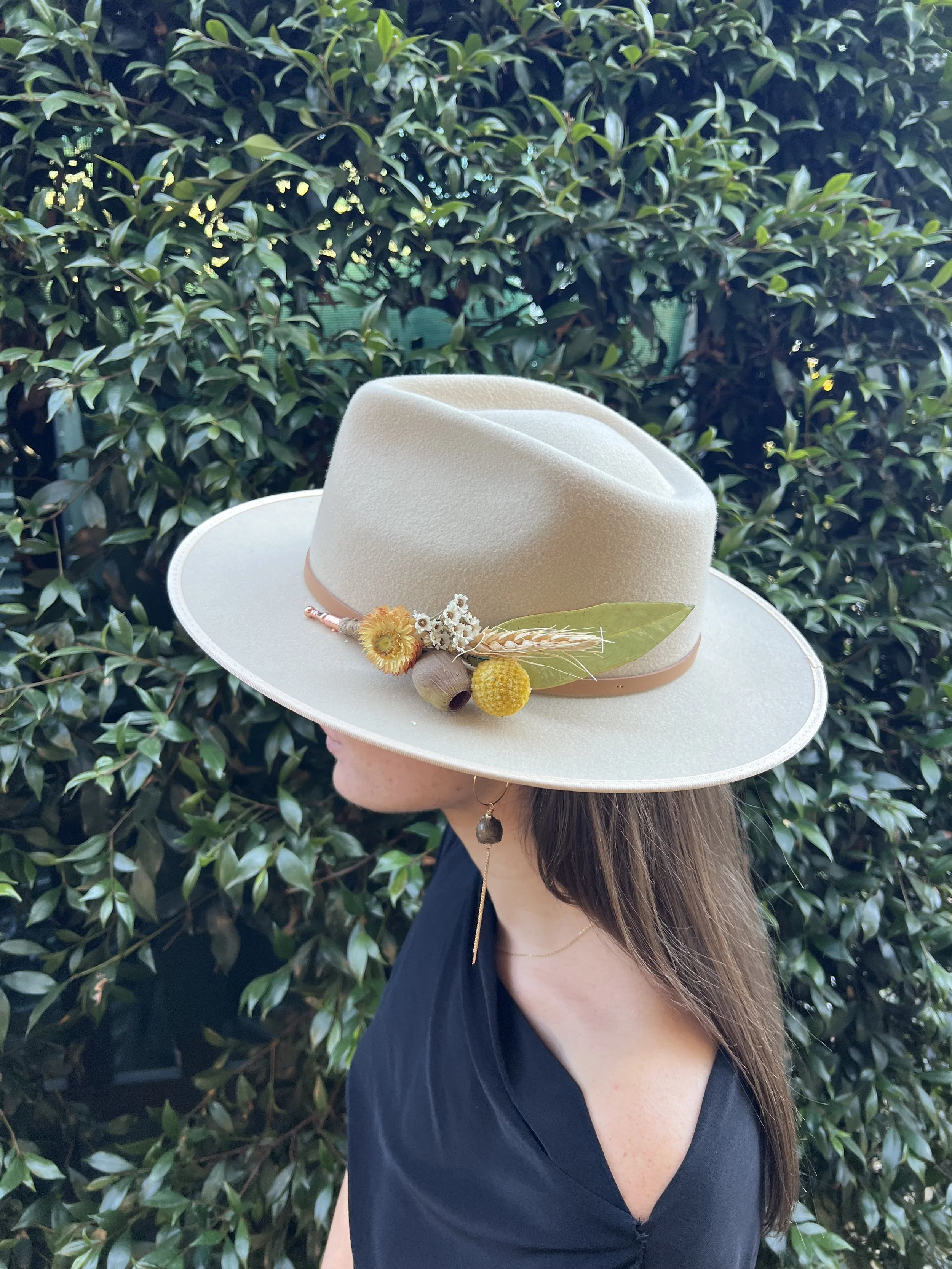 A woman wearing a large beige hat decorated with flowers and leaves, standing outdoors against a leafy green background.
