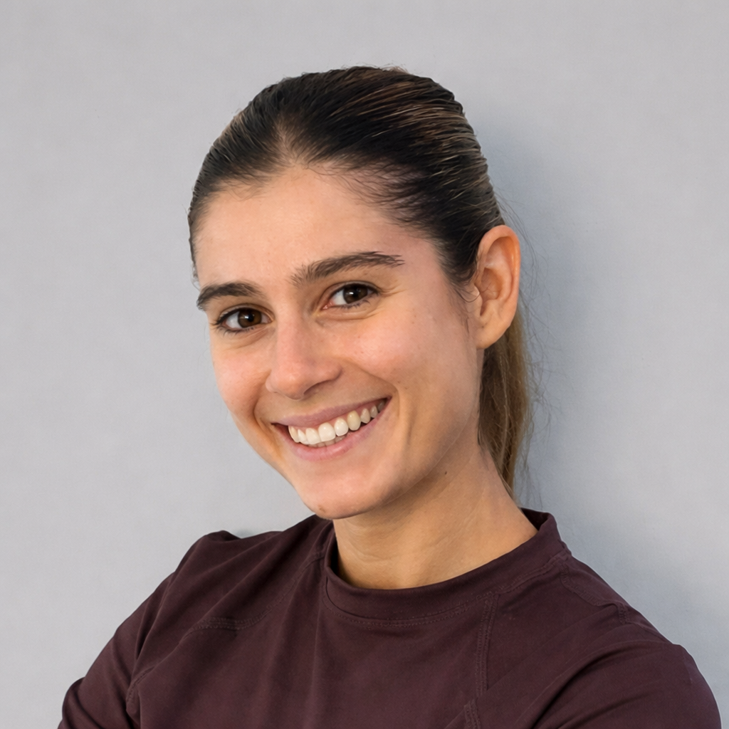 Mujer sonriendo con cabello recogido, usando camiseta marrón oscuro, contra fondo de pared blanca.