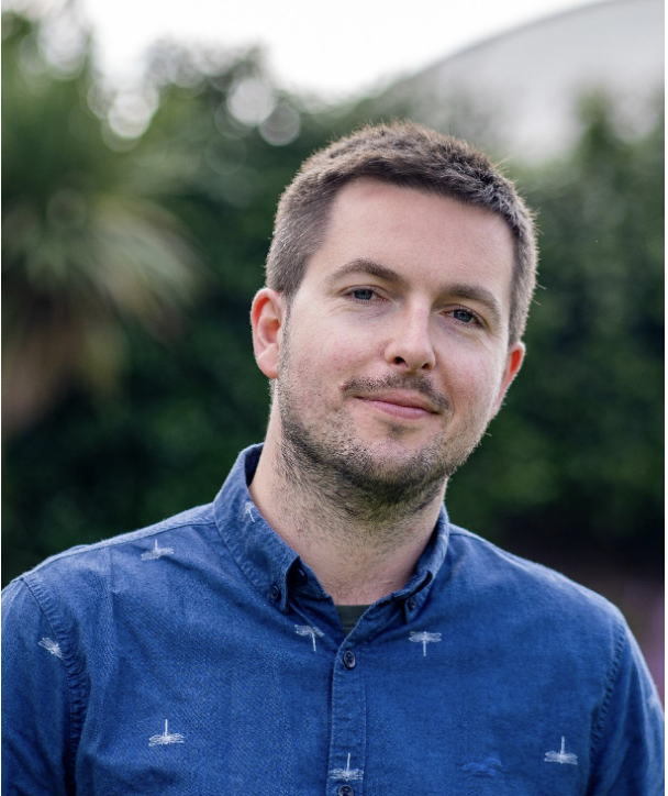 A young man with short brown hair and a slight beard, wearing a blue button-up shirt, standing outdoors with a blurred background of greenery.