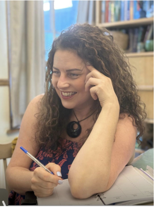 A woman with curly hair smiling and holding a pen, seated at a desk with an open planner, in a cozy room with bookshelves in the background.