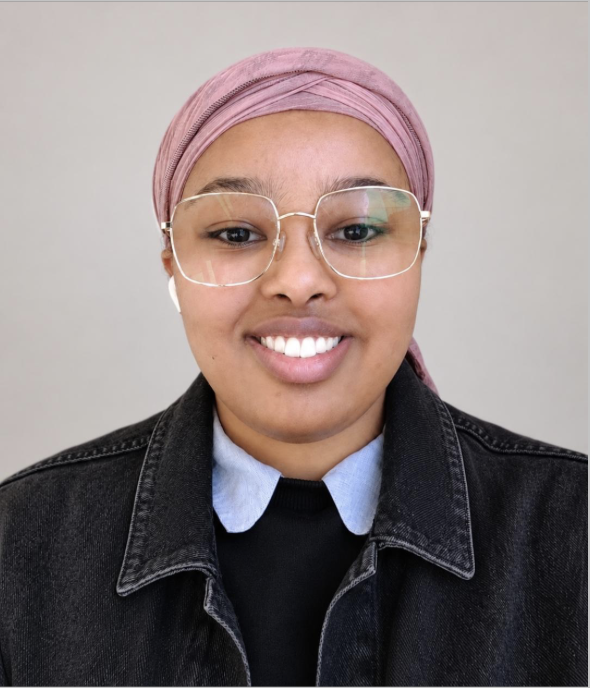 A woman smiling, wearing glasses, a pink headscarf, a black denim jacket, and a collared shirt underneath, against a plain light-colored background.