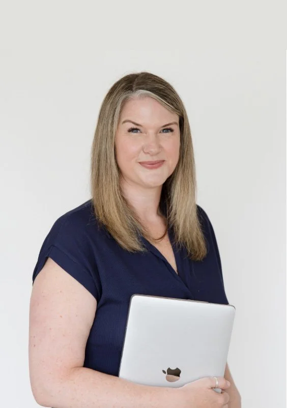 A woman with shoulder-length blonde hair holding a silver MacBook, wearing a navy blue short-sleeve shirt, smiling against a plain white background.