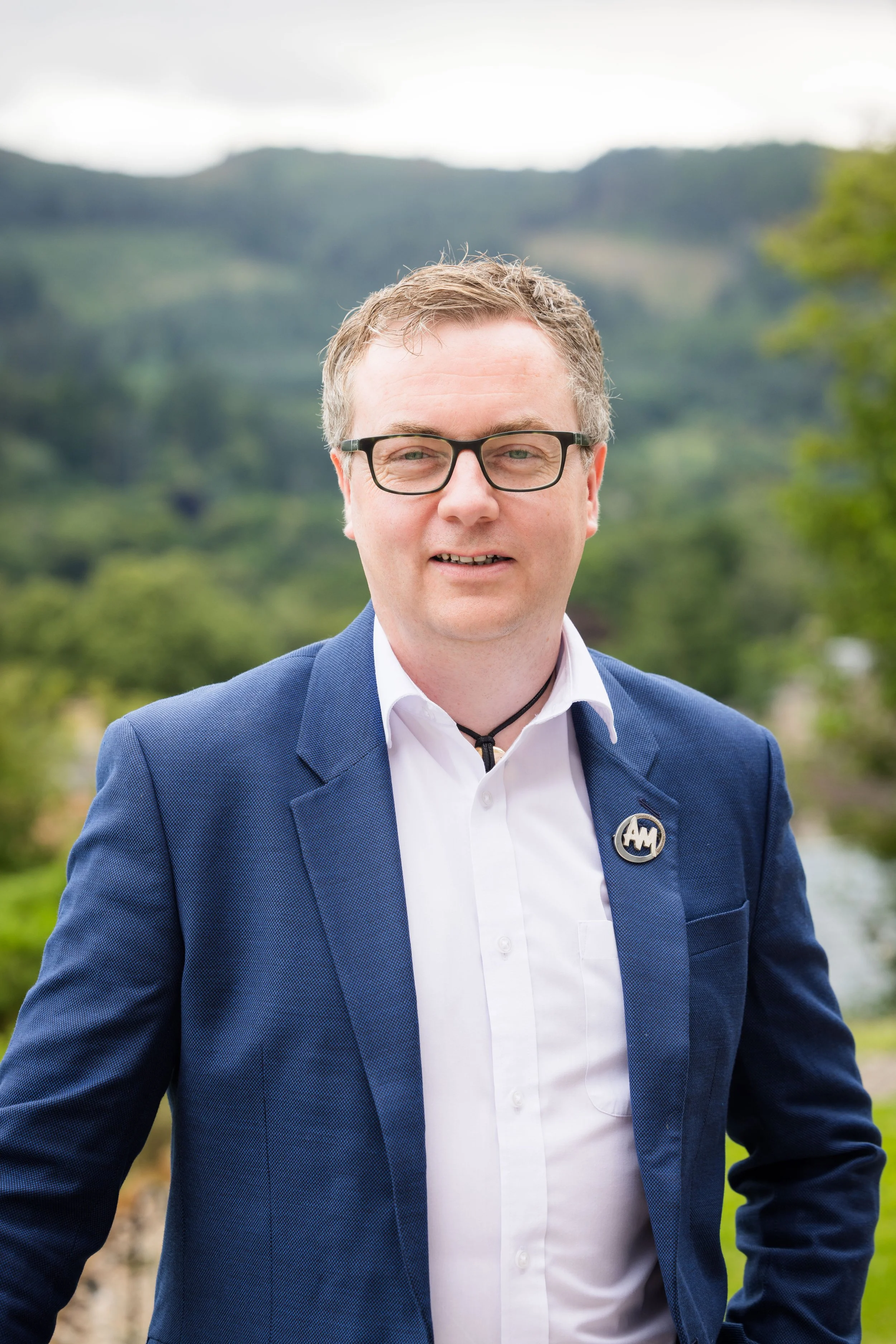 Portrait of a man with glasses wearing a blue suit and white shirt, standing outdoors with a blurred natural landscape background.