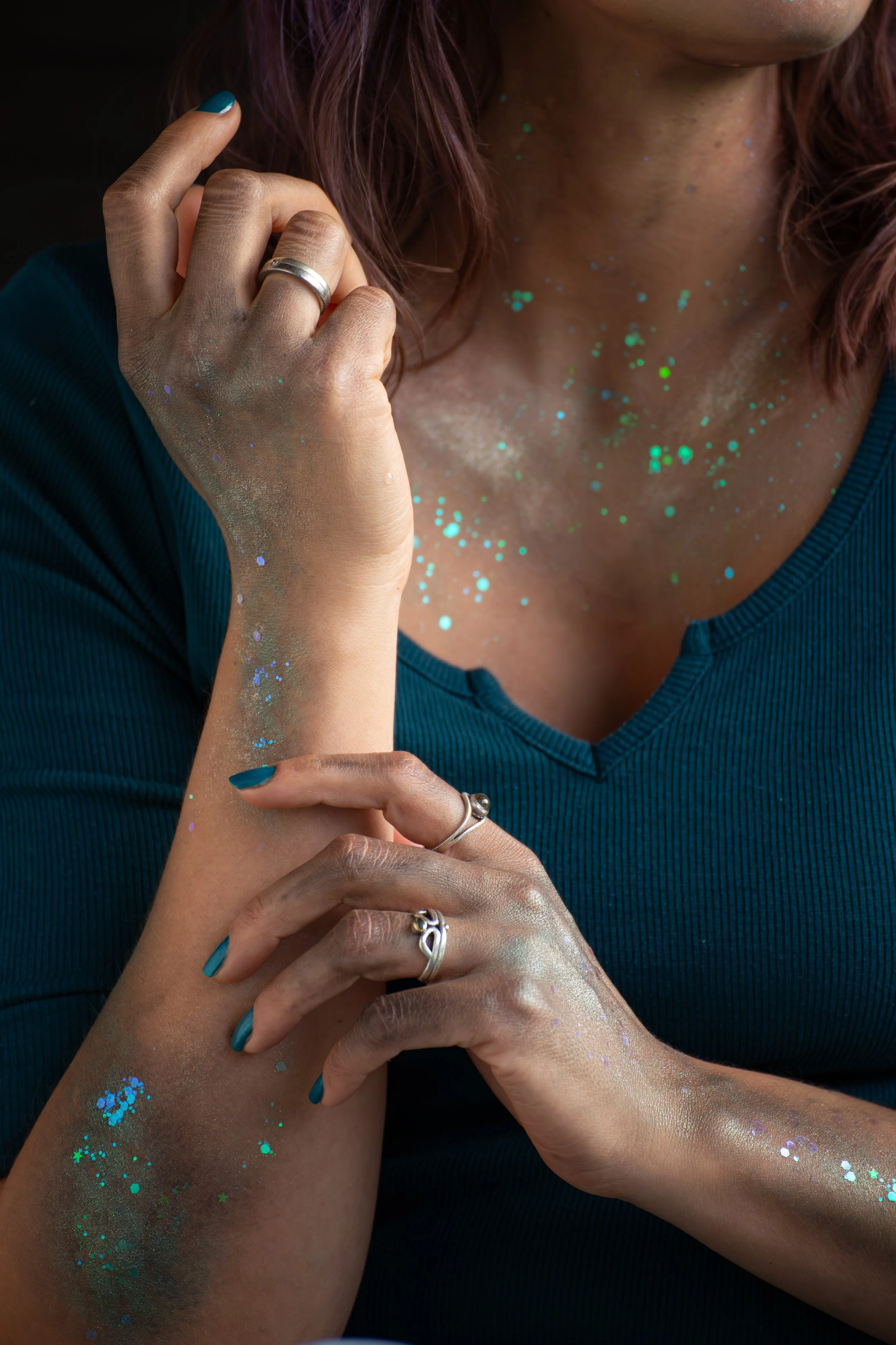 Nahaufnahme einer schönen, zauberhaften Frau mit glitzernder Haut und Fingernägeln, die mit dunklerem Türkis lackiert sind, die Person trägt mehrere Ringe, dunkle Haare und trägt ein dunkelblaues shirt.