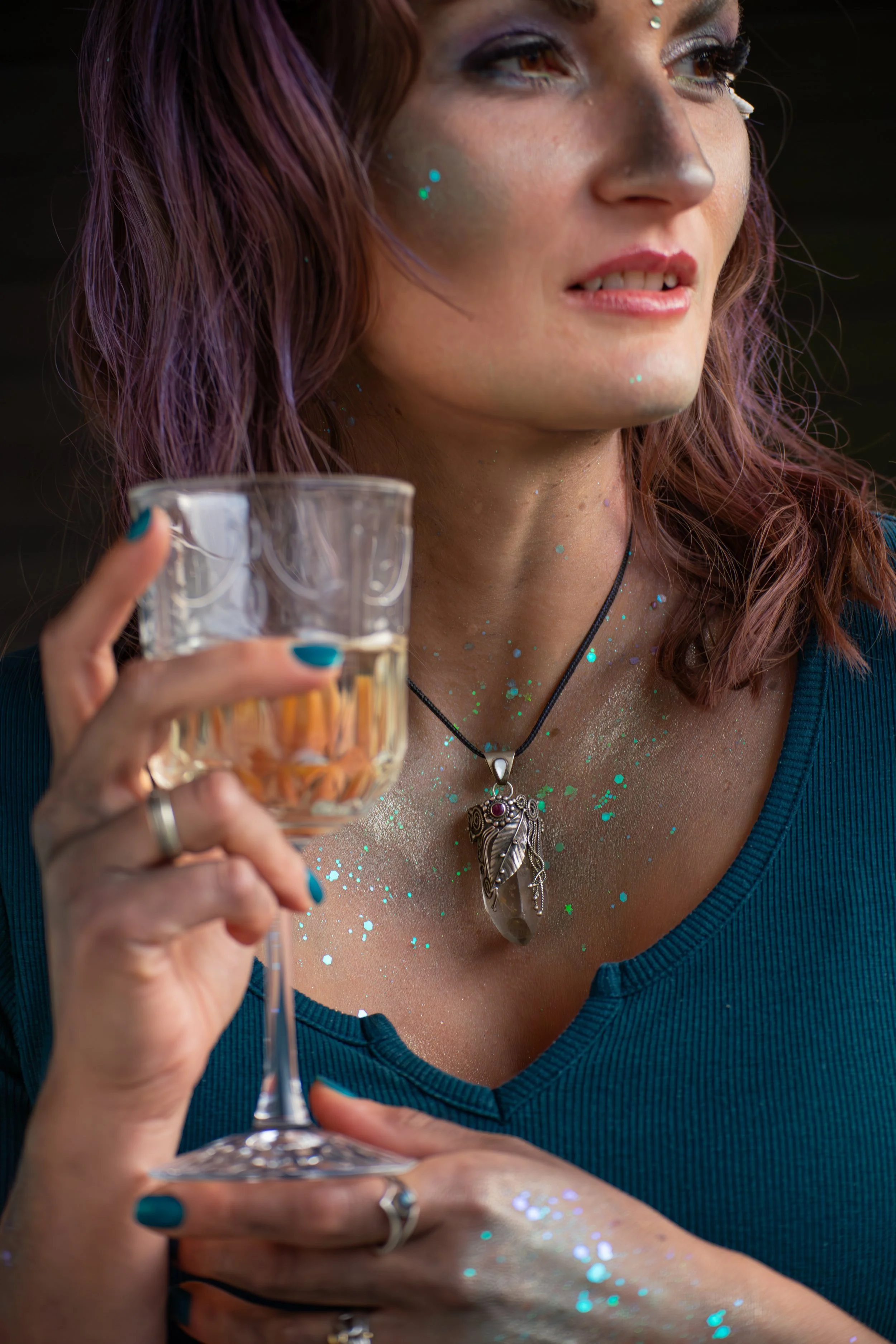 Nahaufnahme einer Frau mit lila Haar, die ein Weinglas hält, trägt einen silbernen Anhänger und trägt blauen Top, mit Glitzer auf Haut und Hals.