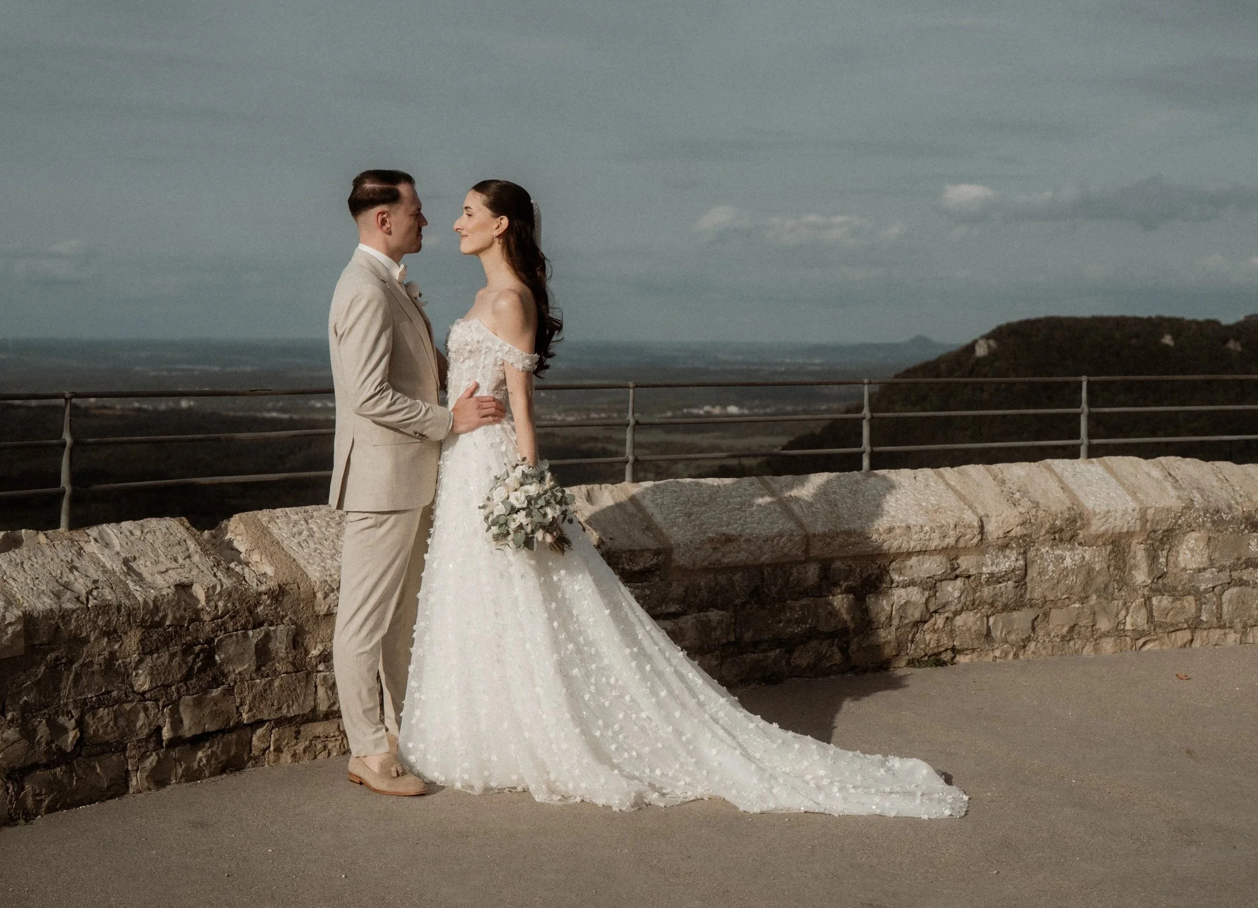 Ein Brautpaar steht sich auf einer offenen Terrasse gegenüber, im Hintergrund ein Landschaftshintergrund mit Bergen und Himmel, sie sind in Hochzeitskleidung, die Braut hält einen Blumenstrauß.