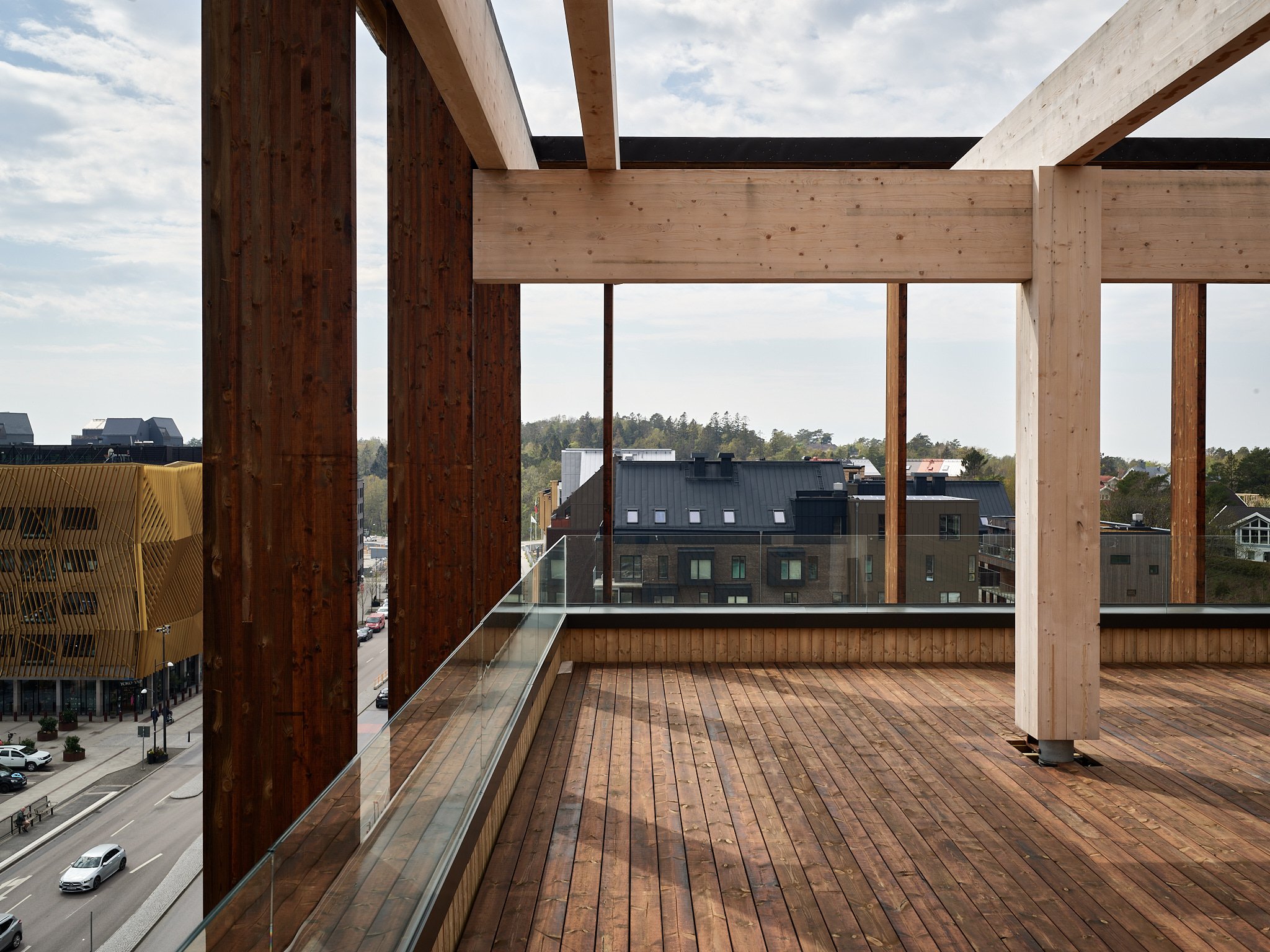 View from a building balcony with glass railing, showing a street and neighboring buildings, with wooden beams and deck, under a partly cloudy sky.