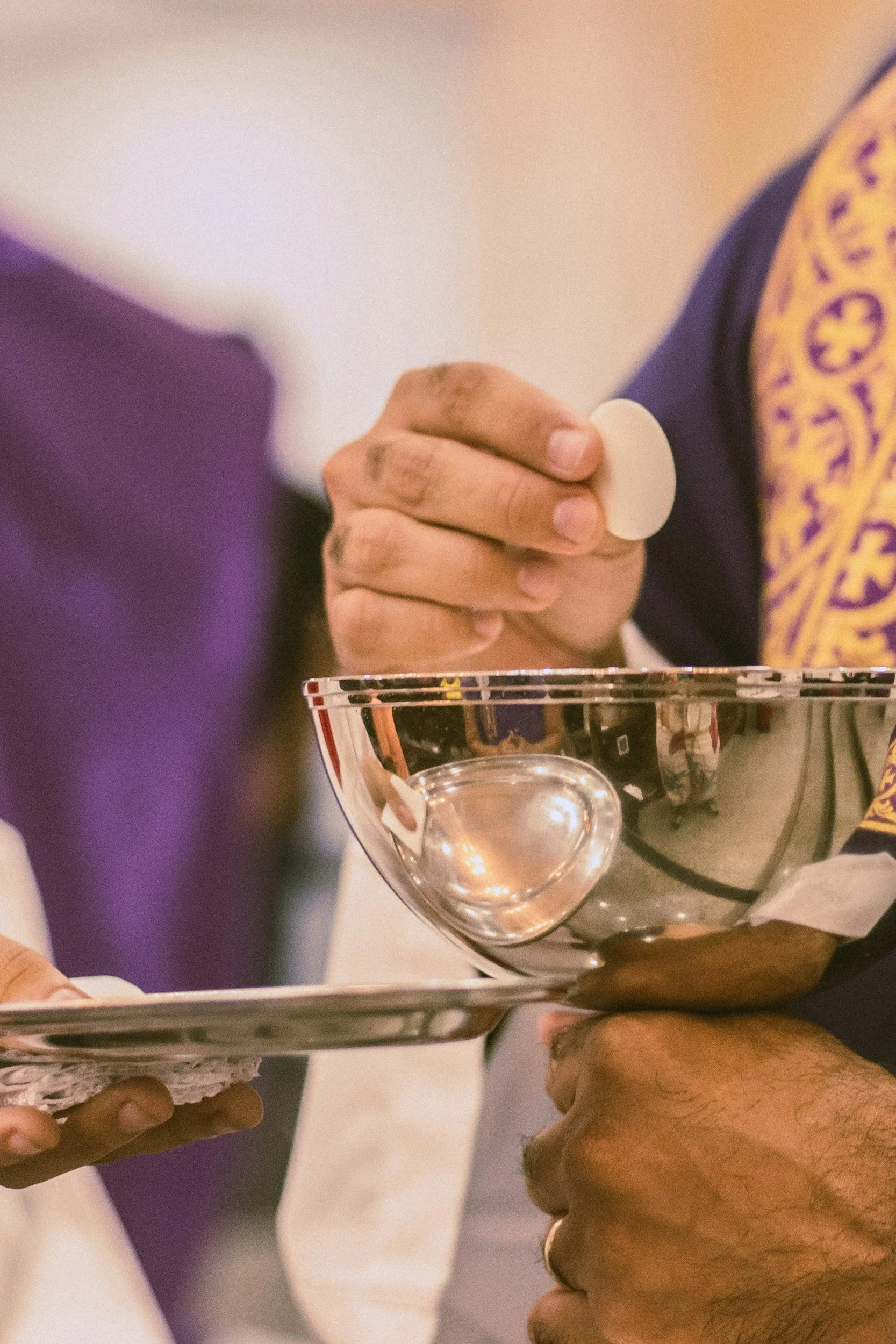 A priest holding a communion in one hand and reaching towards chalice with the other hand. The priest is wearing purple and gold.