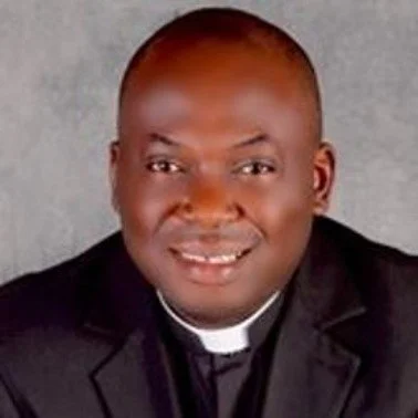 Portrait of a man wearing a black suit and clerical collar against a gray background.