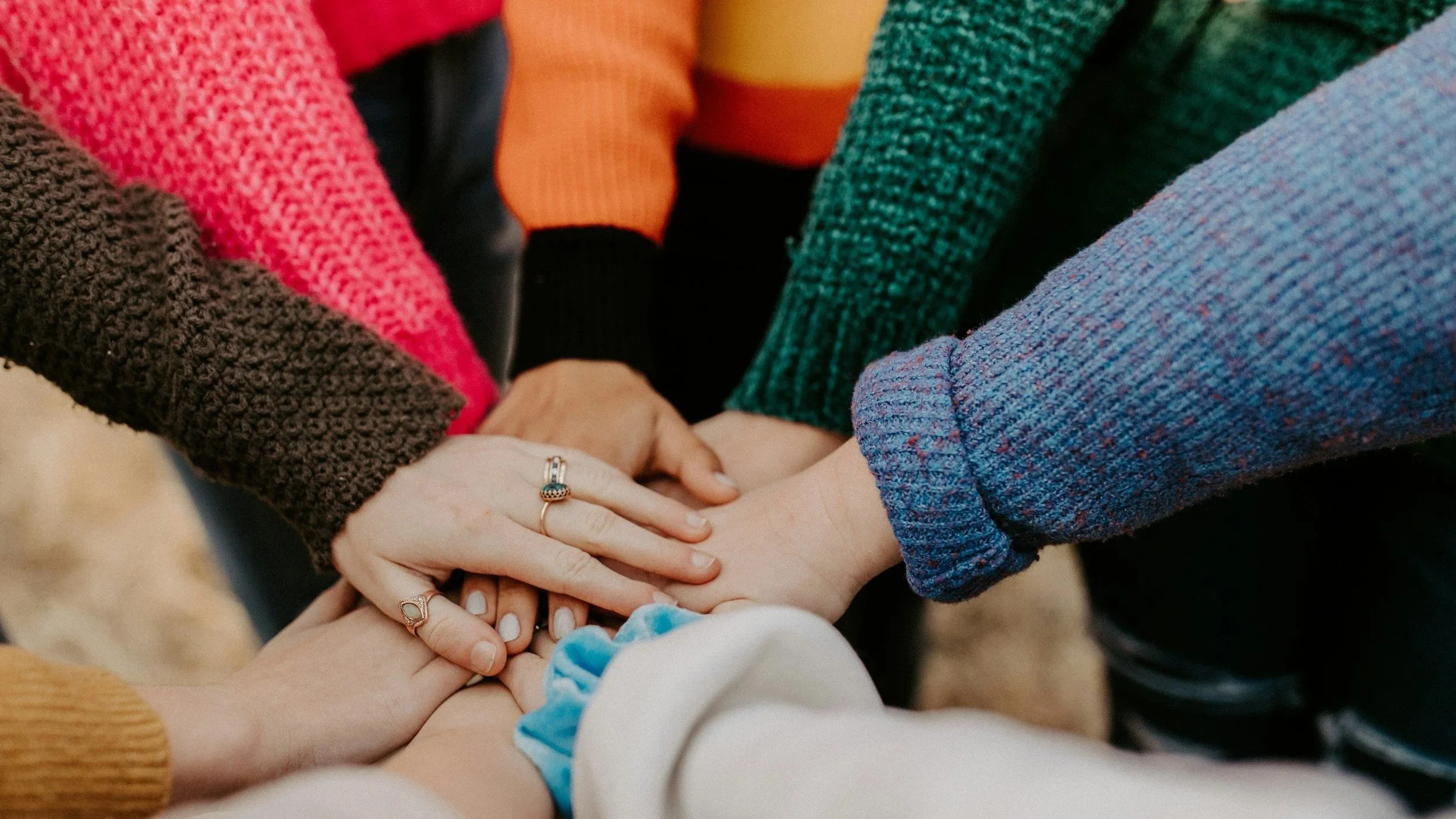 Group of people holding hands together in a unity gesture, focusing on their hands and colorful sweaters.