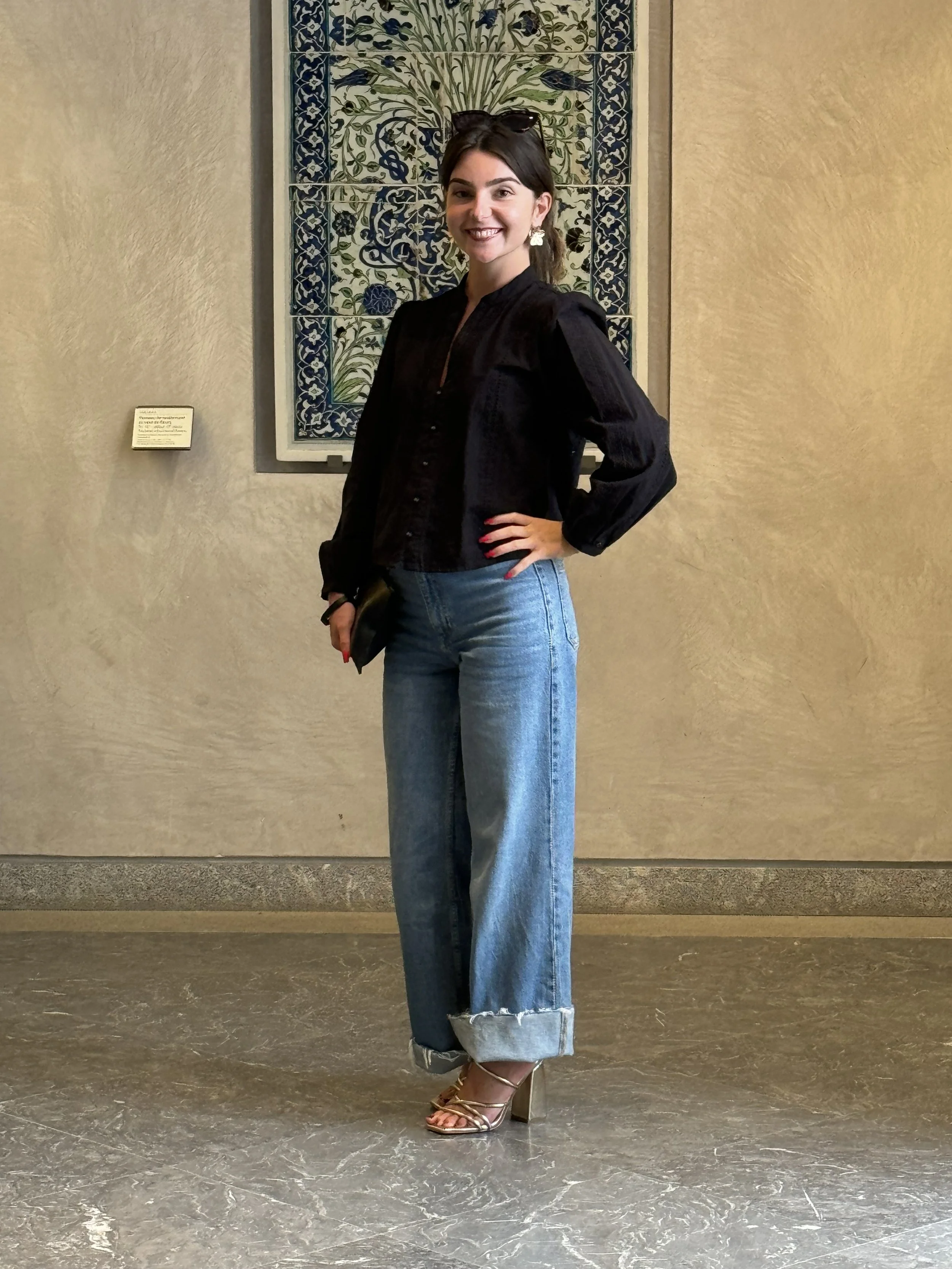 Une femme souriante pose dans un musée ou une galerie d'art, portant une veste noire, un jean mom à revers, des talons dorés et des lunettes sur la tête, avec une œuvre d'art en mosaïque de fleurs en arrière-plan.