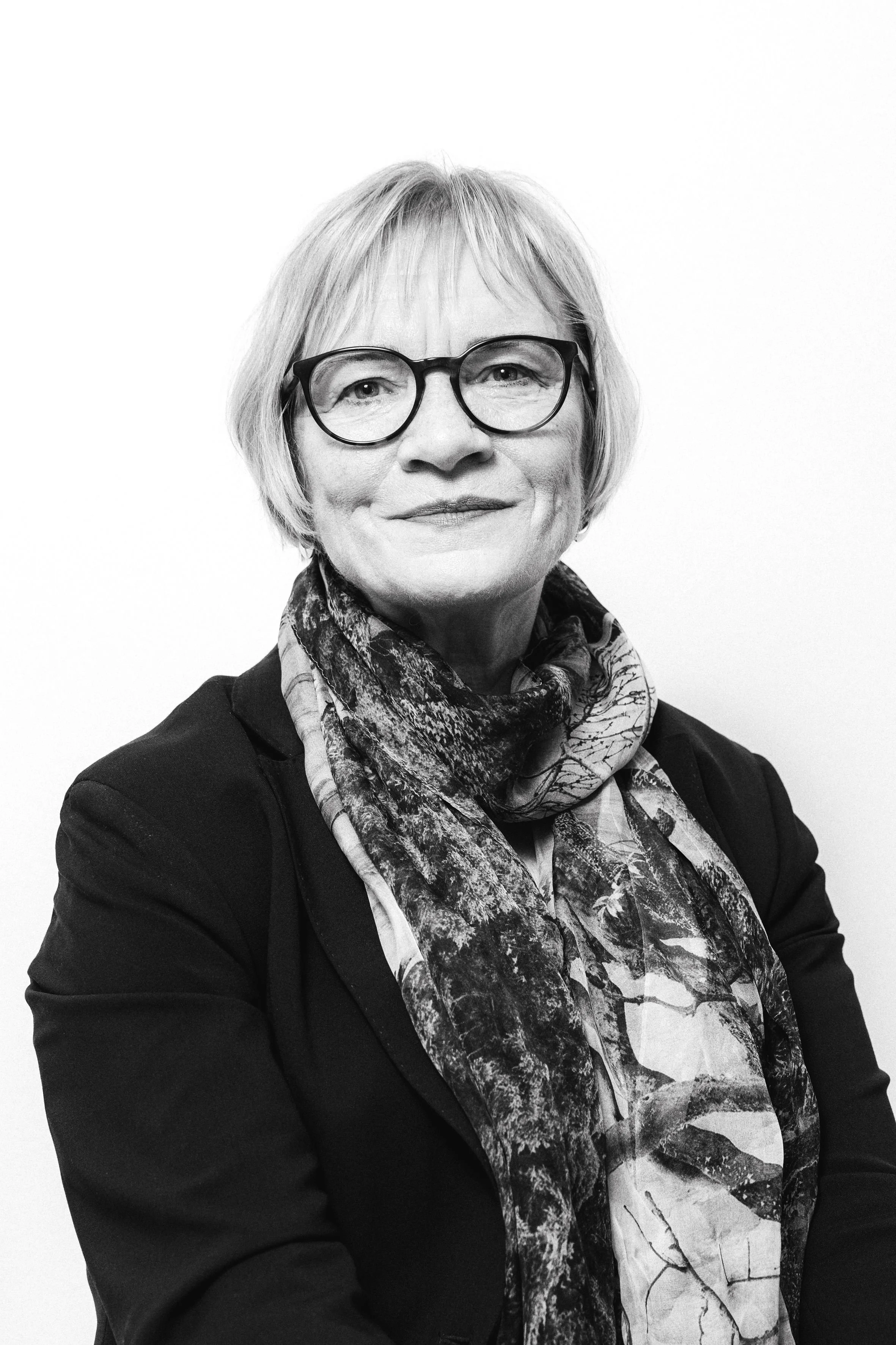 A black-and-white portrait of a middle-aged woman with short, straight hair, wearing glasses with round frames, against a dark background.