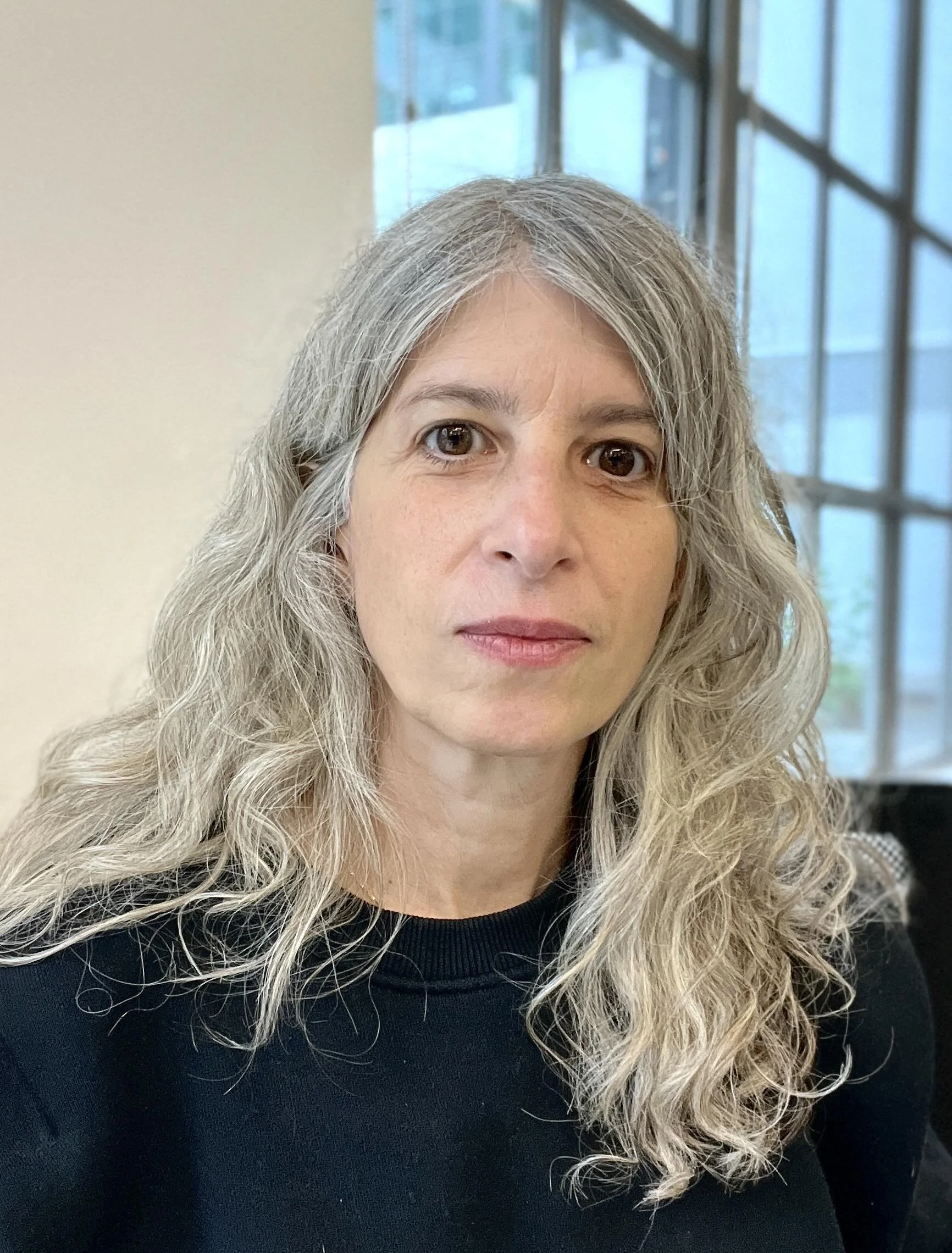 A woman with gray, wavy hair and fair skin, wearing a black top, sitting indoors near a window with a grid of panes, with a neutral facial expression.