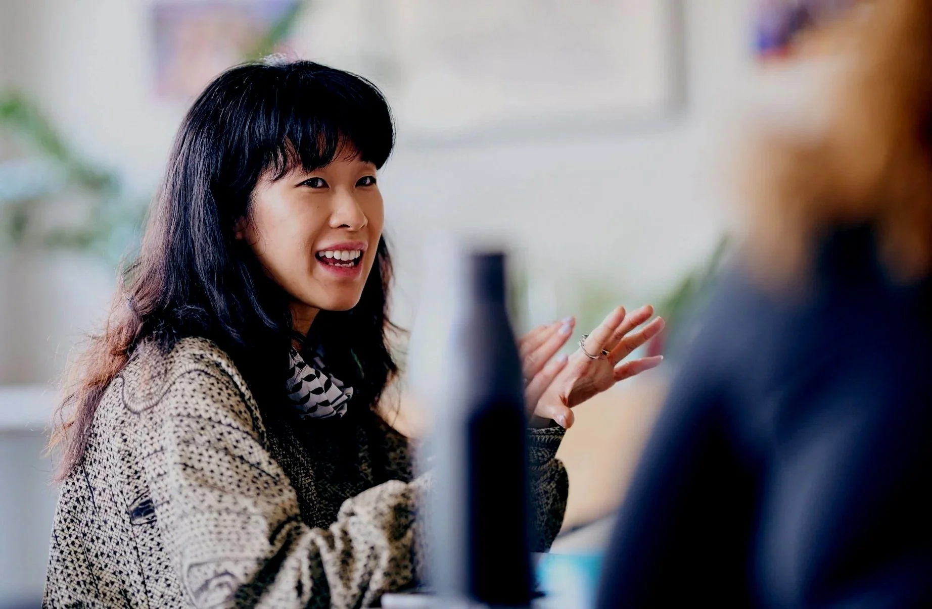 Young woman with long black hair and a patterned sweater smiling and gesturing during a conversation in an indoor setting.
