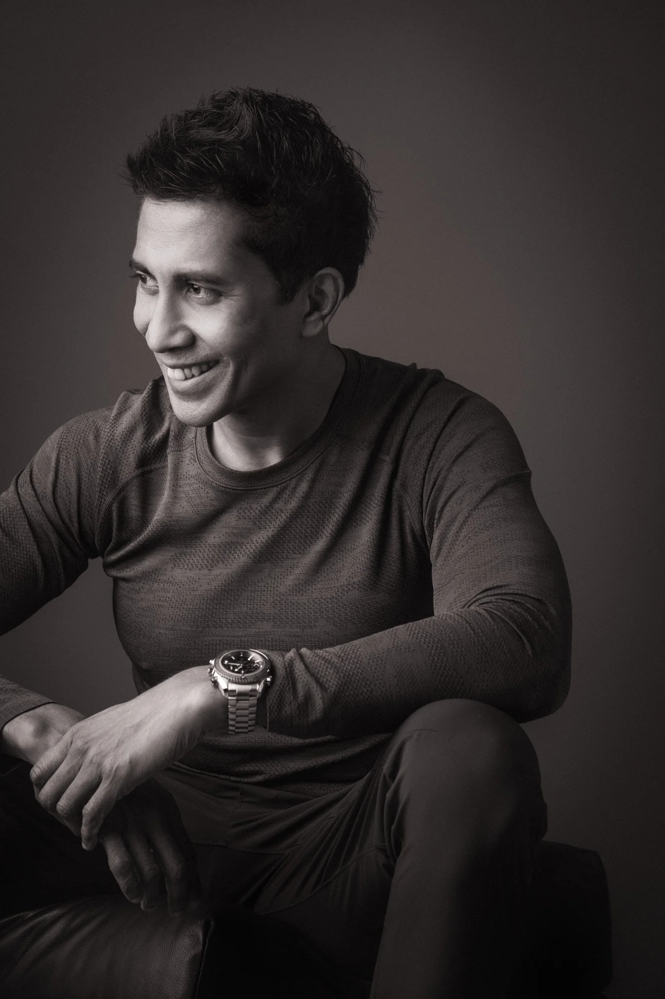 Black and white photo of a young man with dark, wavy hair smiling, wearing a watch, a long-sleeved shirt, and dark pants, sitting on a leather chair against a plain background.