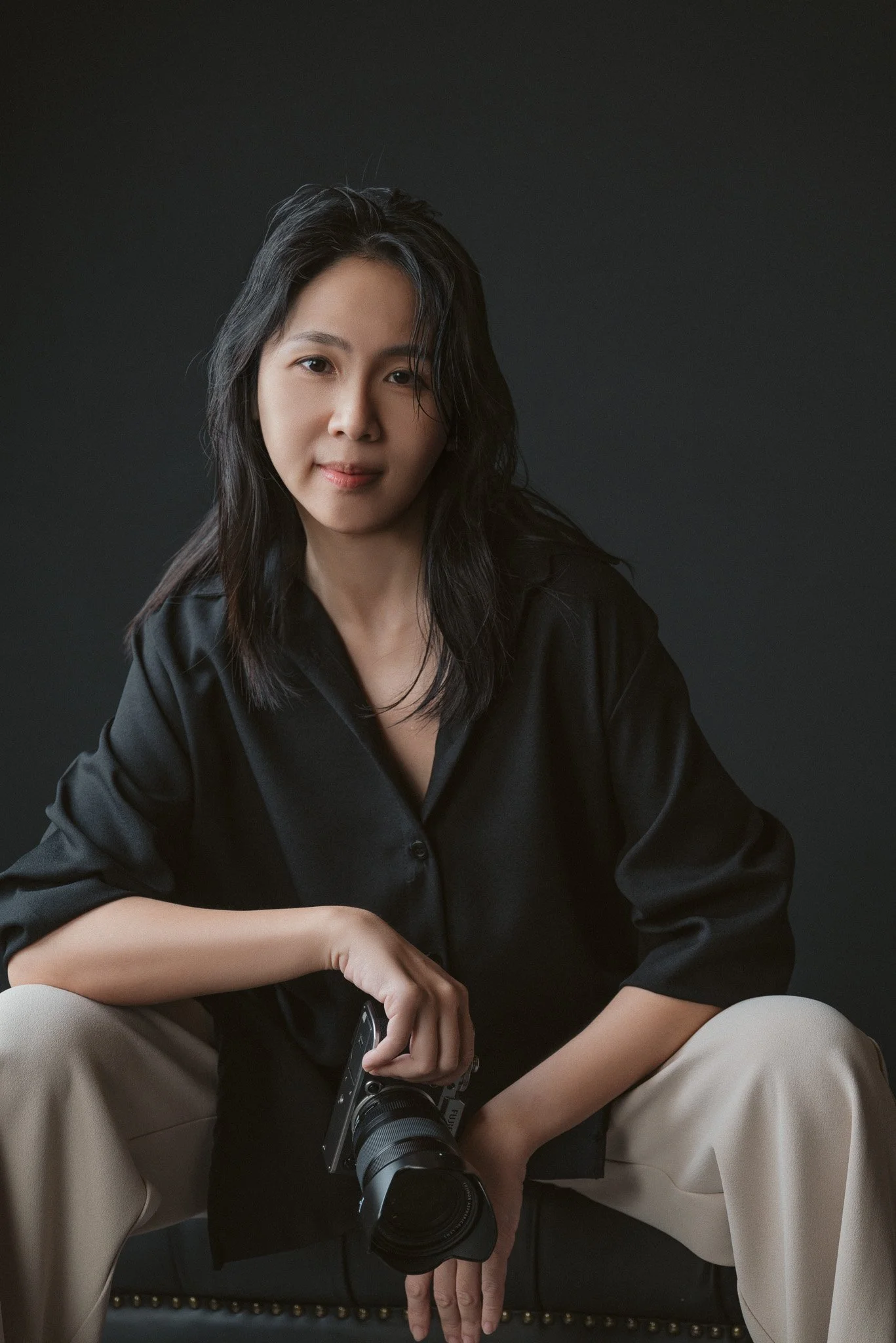 A woman sitting on a black chair, wearing a black shirt and beige pants, holding a camera with a large lens. She has long, dark, wavy hair and is looking at the camera in front of a dark background.
