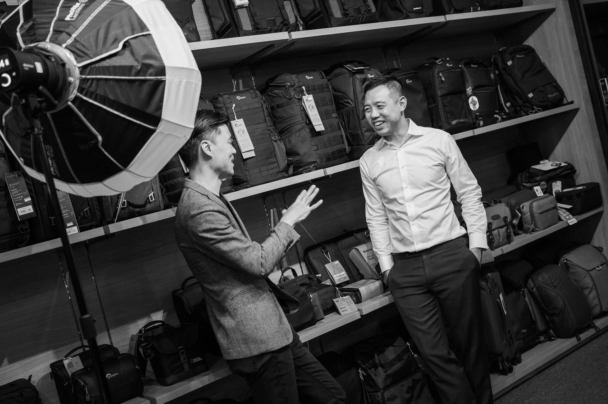 Two men talking in a camera store surrounded by camera bags on shelves.