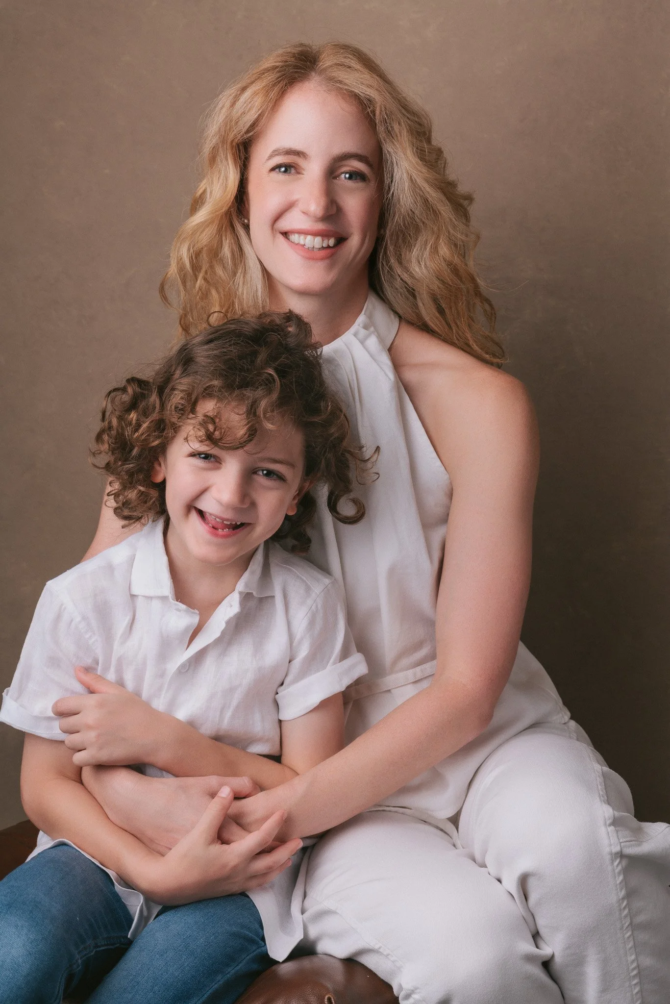 A woman with blonde hair smiling and sitting, holding a young boy with curly brown hair and a big smile, both dressed in white shirts, against a neutral background.