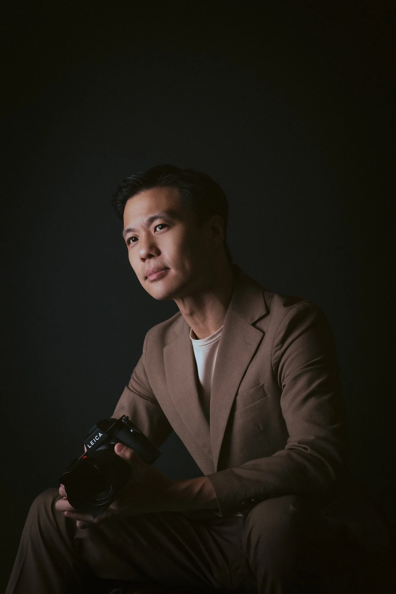 A man dressed in a brown suit and white shirt, holding a Leica camera, sitting against a dark background.