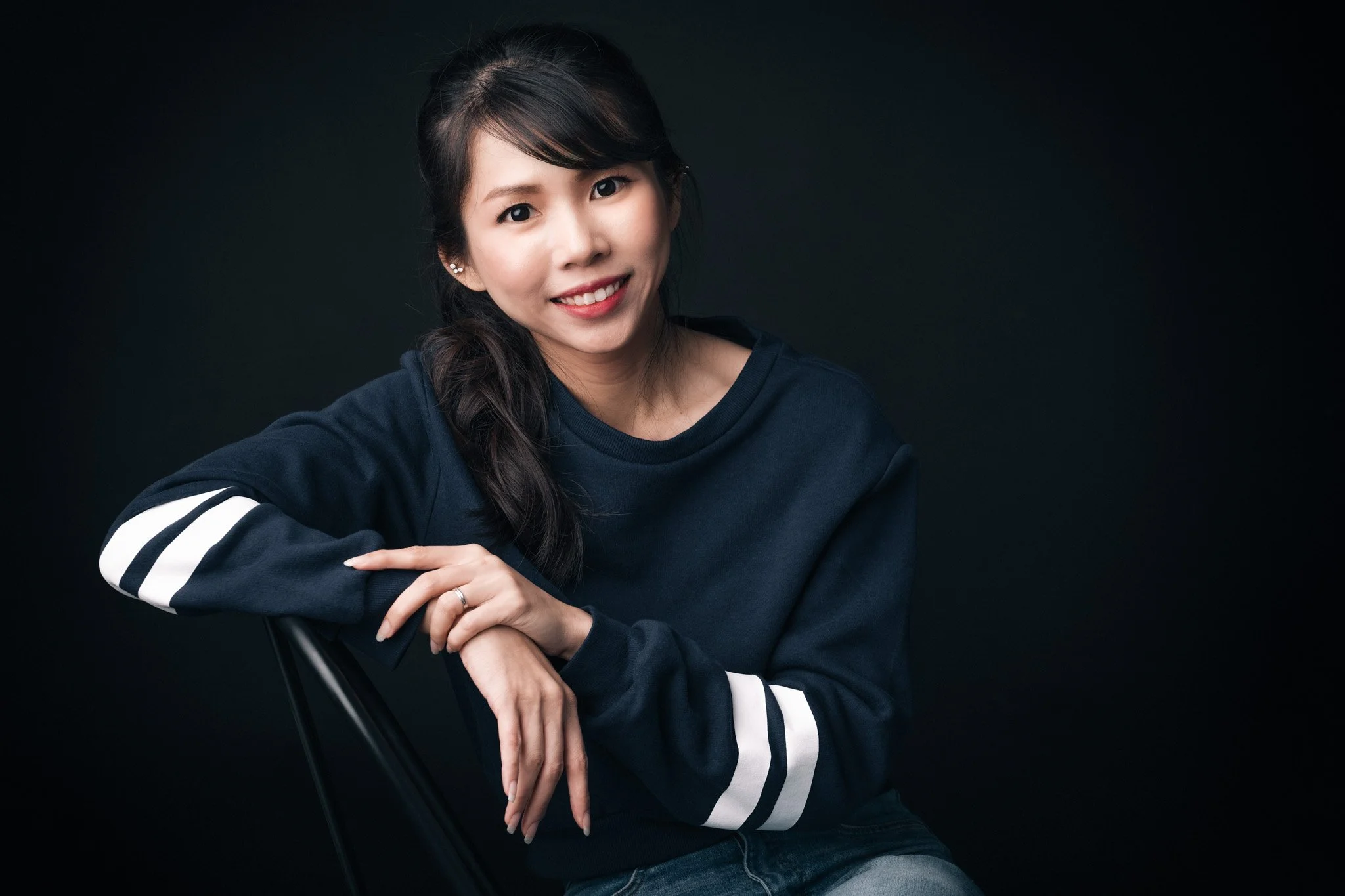 A young woman with long dark hair and earrings smiling while sitting against a black background, wearing a dark sweatshirt with white stripes on the sleeves.