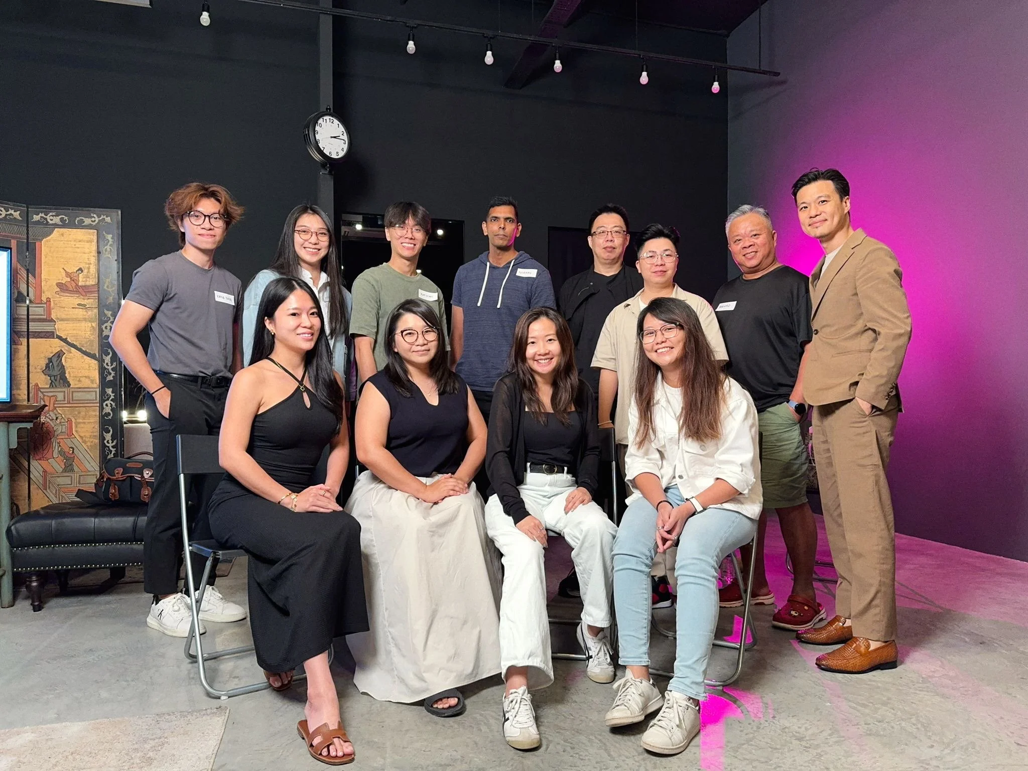 A diverse group of eleven people, including men and women, is posing together in an indoor space with black and purple walls, with some seated and others standing, smiling at the camera.
