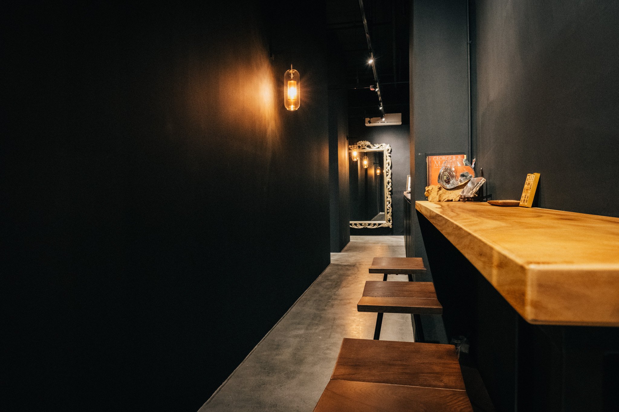 A dimly lit modern interior with a long wooden bar counter, hanging light fixtures, and a decorative mirror at the end of the corridor.
