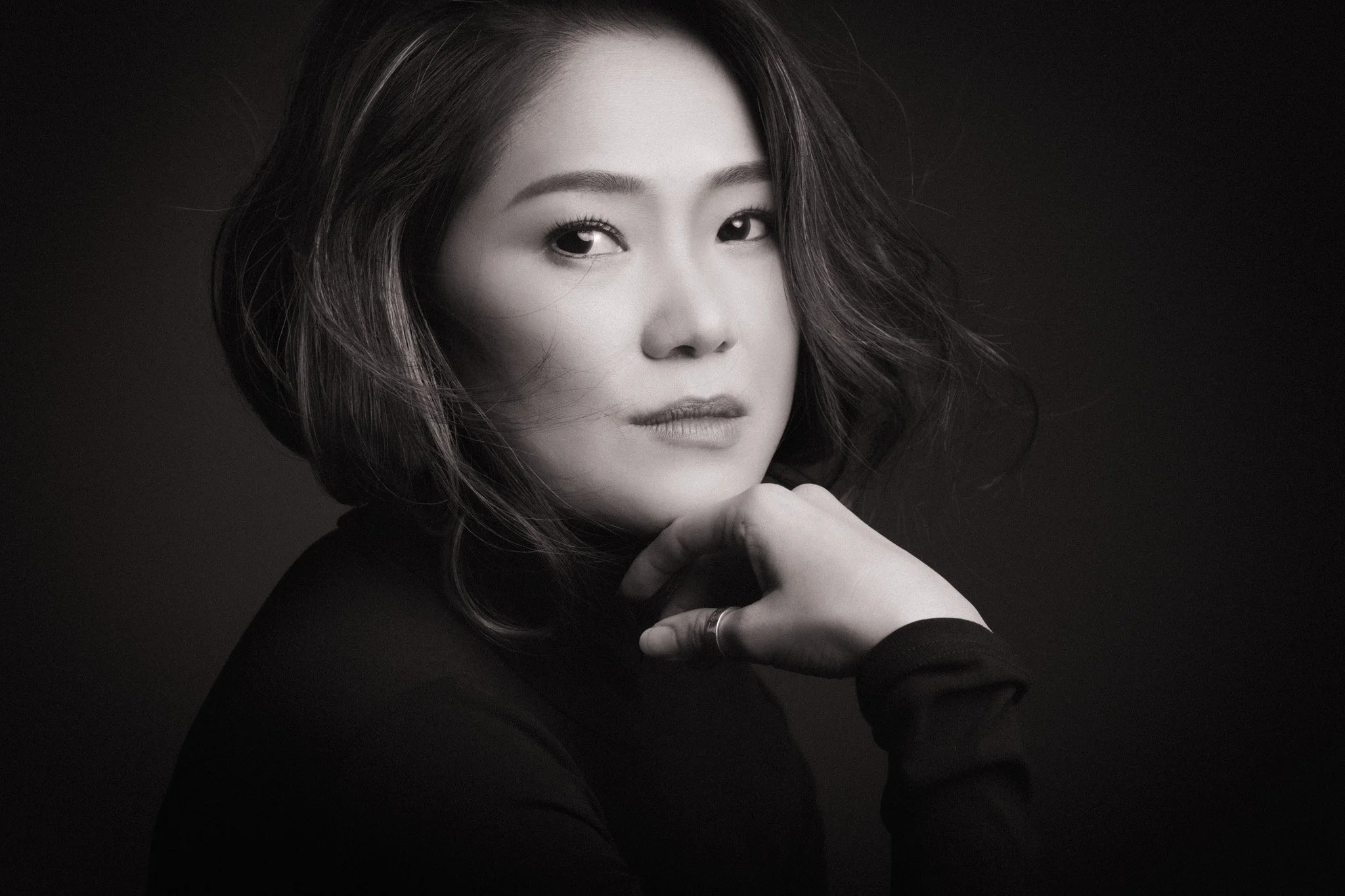 Black and white portrait of an Asian woman with shoulder-length hair, gazing softly into the camera, with her hand resting near her face.