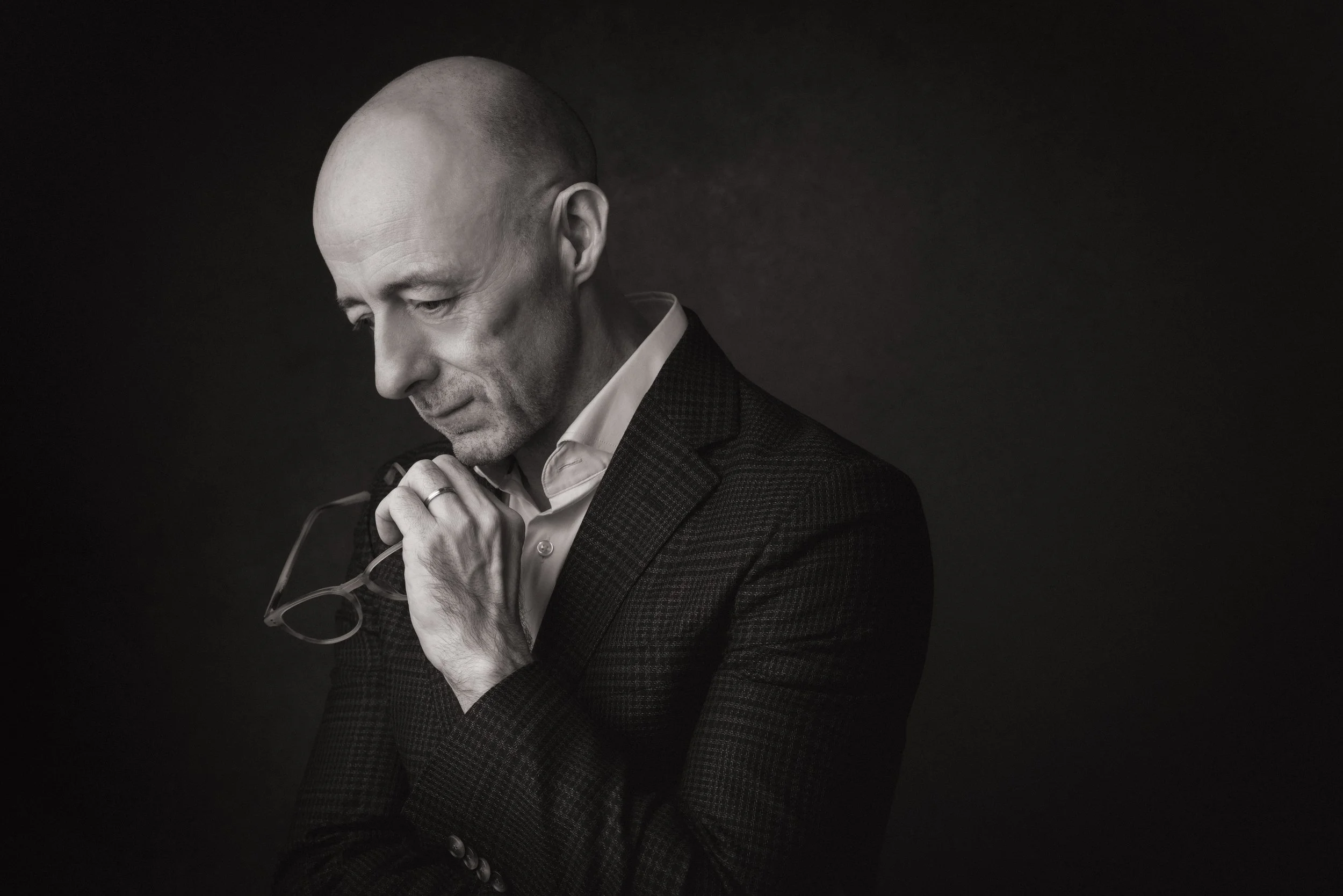 A black and white portrait of a bald man wearing a suit, holding glasses, with his head slightly bowed and eyes closed.