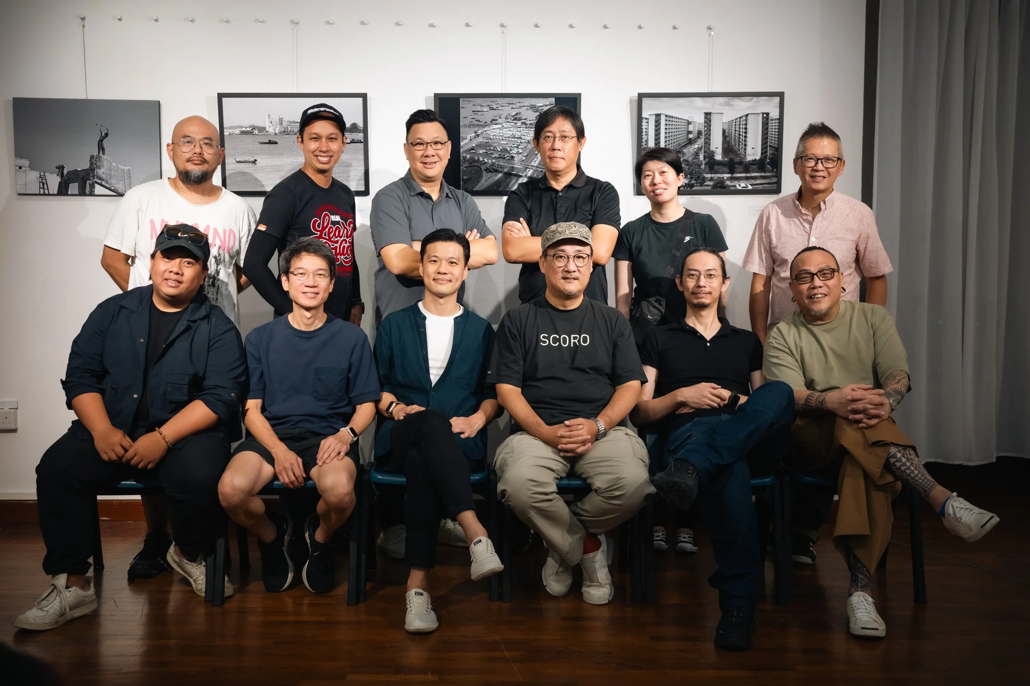 A group of fifteen diverse people, some sitting and some standing, posing for a photo in front of black-and-white framed photographs on a white wall. The group includes men and women of various ages, some wearing glasses, casual clothing, and sneakers, with a wooden floor beneath them.
