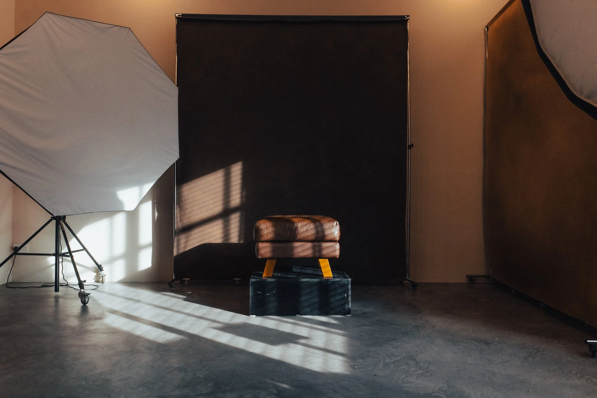 Photography studio setup with a large softbox light on the left, a black backdrop in the center, and a brown padded stool in front of the backdrop. The lighting creates shadows on the plain concrete floor.