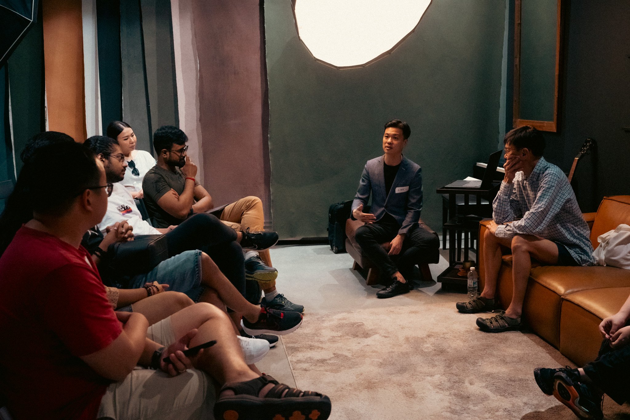 A group of people sitting in a circle in a cozy room, engaging in a discussion or workshop with a man speaking. The room has soft lighting and a green and brown textured wall with a large round light fixture overhead.