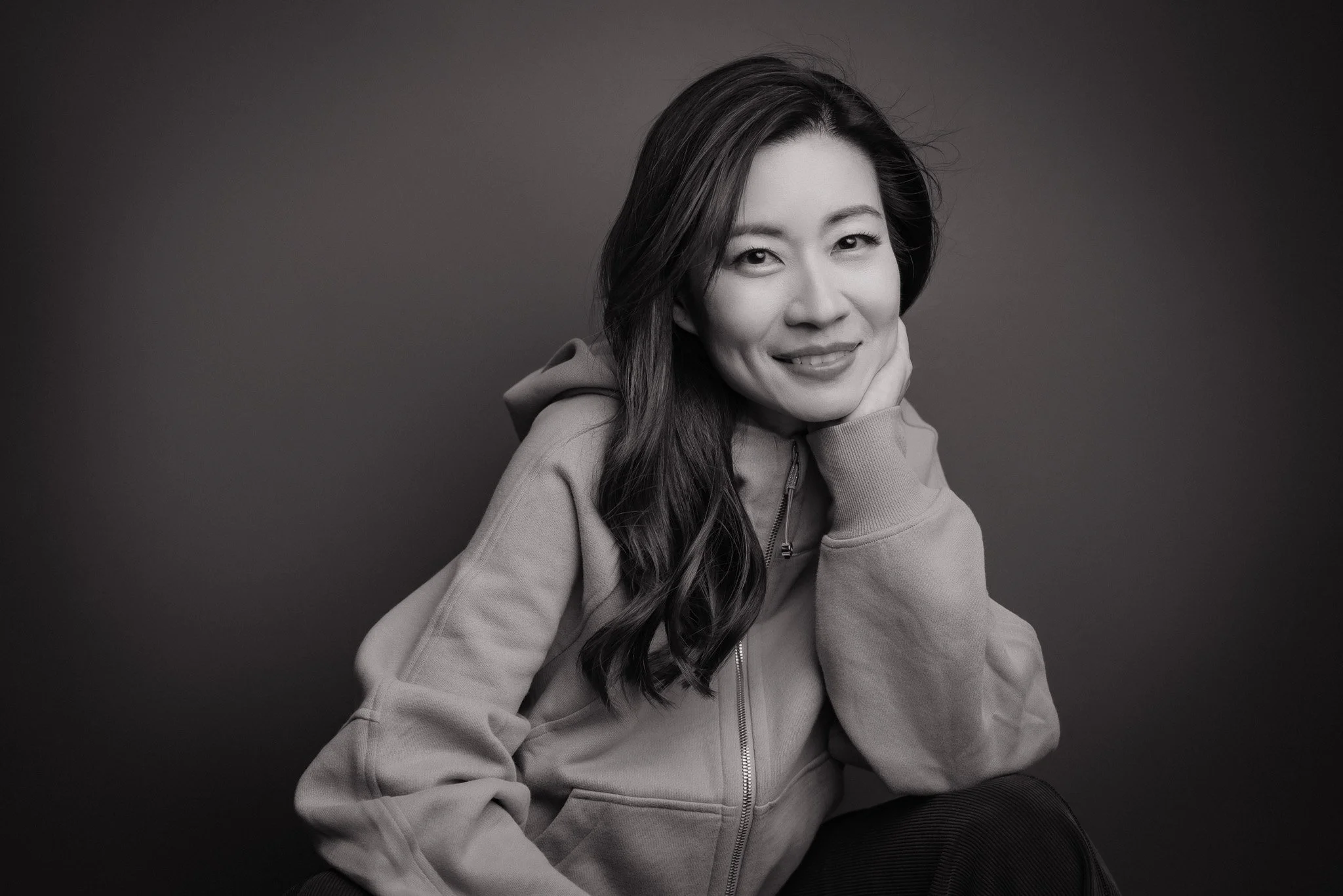 Black and white portrait of a smiling woman with long hair, wearing a hoodie, seated against a plain background, looking at the camera.