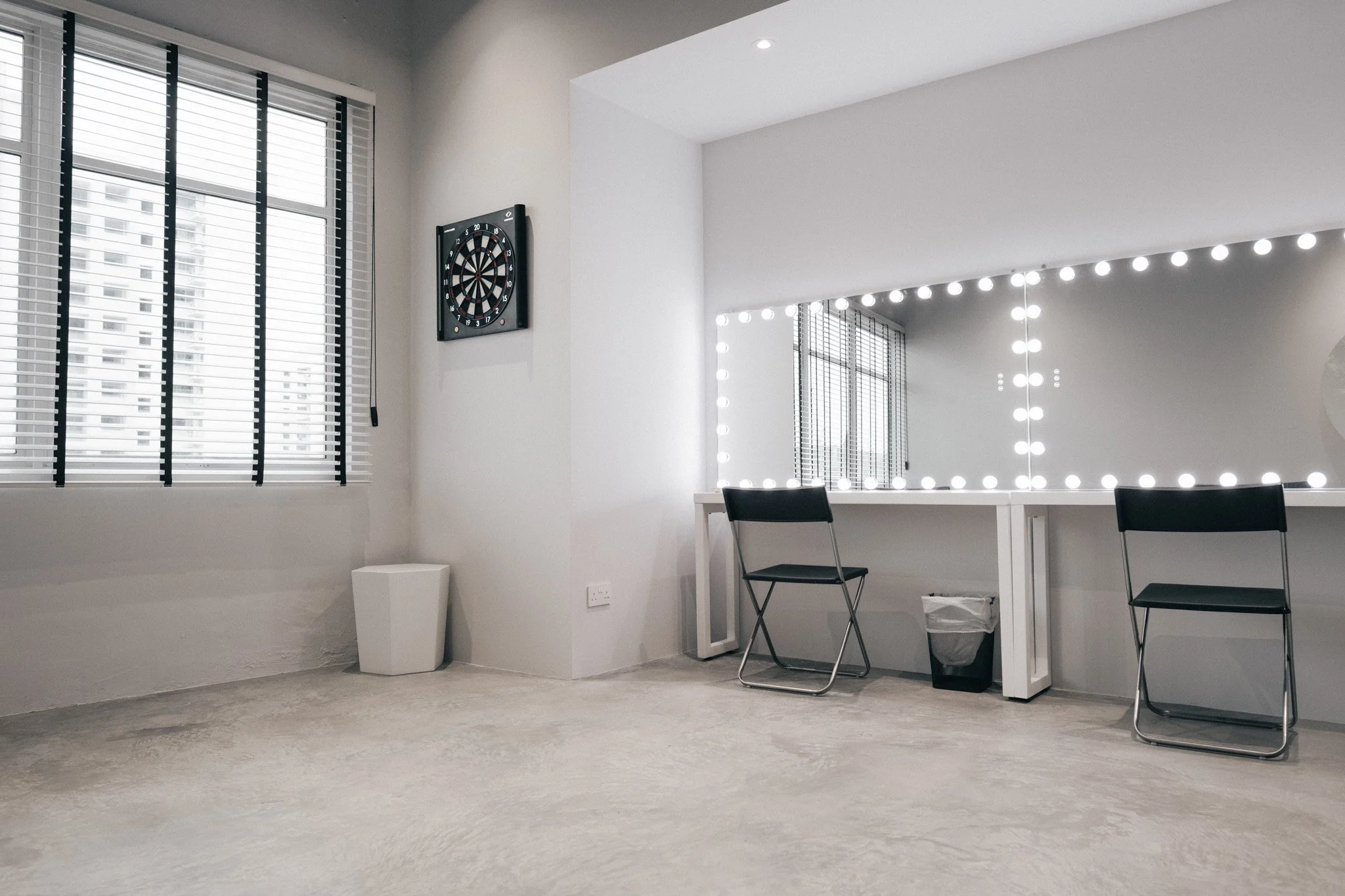 Empty room with large backlit mirror, black chairs, dartboard on wall, and window with blinds.