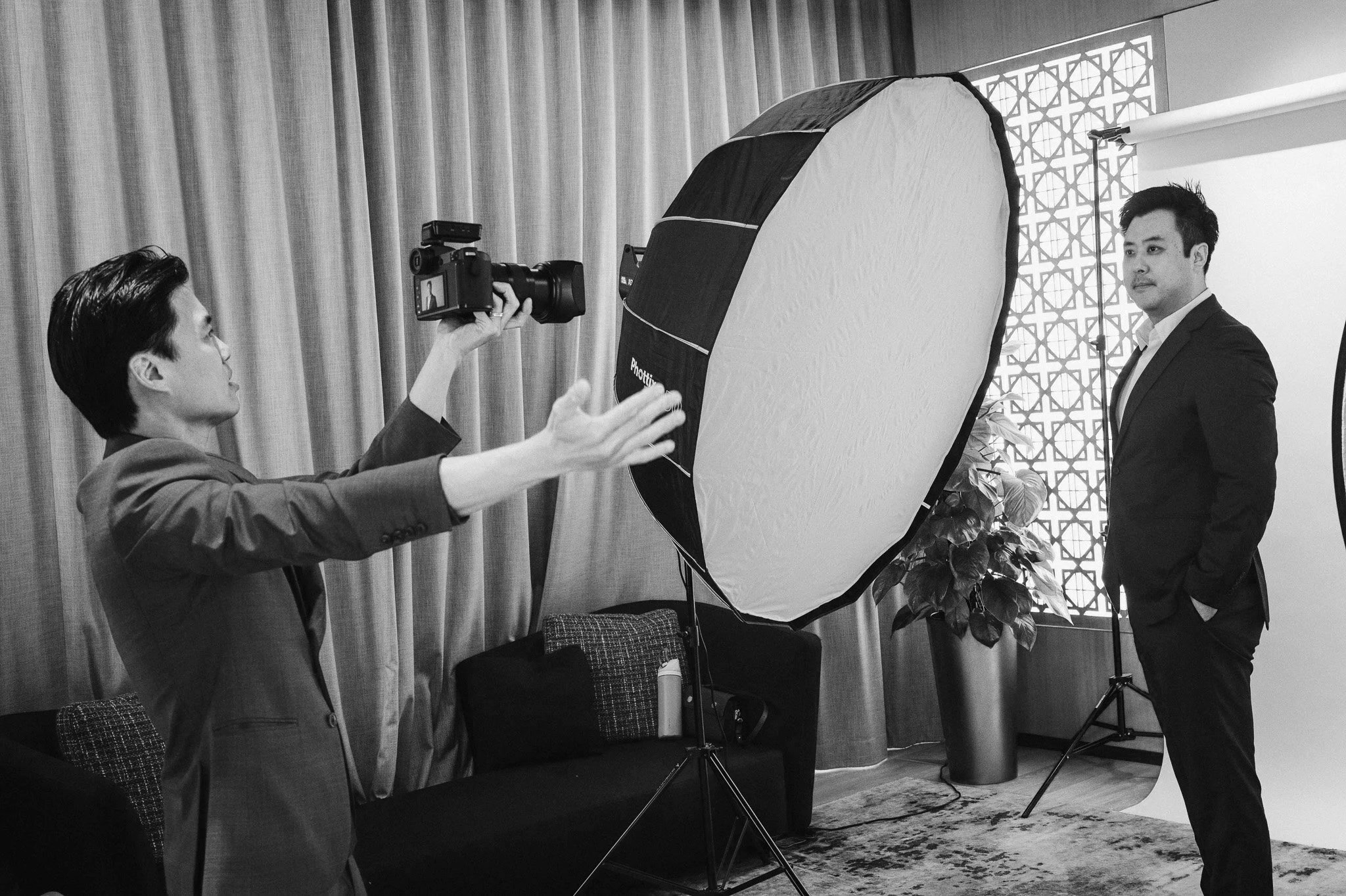 A man in a suit standing in front of a camera and a softbox, preparing for a photoshoot in a studio with curtains and decorative windows.