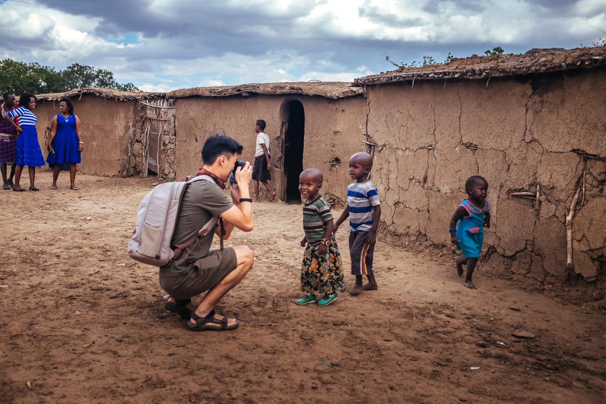 A man kneeling on the ground taking a photo of three young children walking in front of a mud house. Several women stand nearby, watching and smiling. The scene is outdoors with cloudy skies.