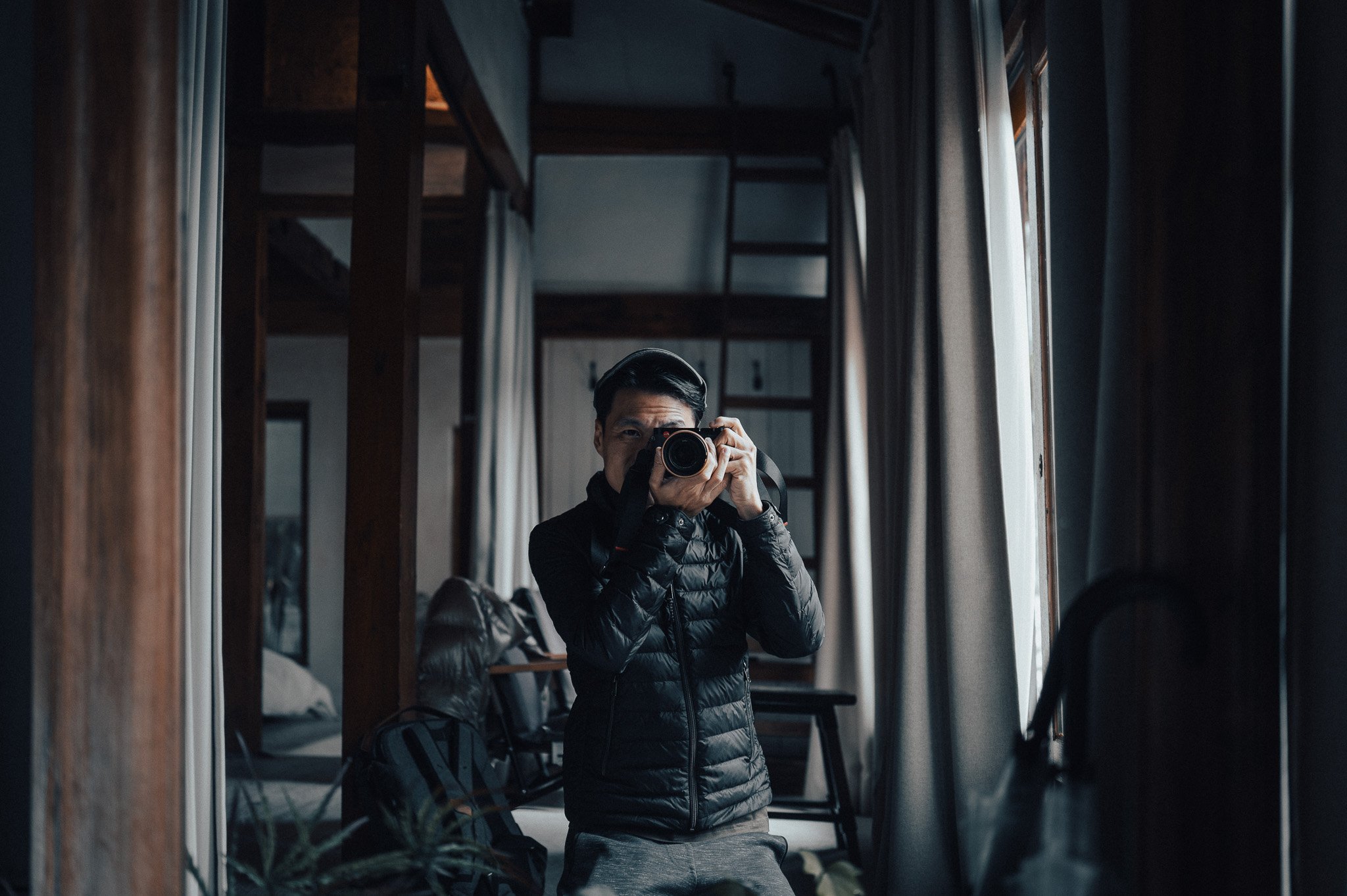 A man taking a photo of himself in a mirror with a camera, wearing a black jacket, in a room with wooden beams and windows.