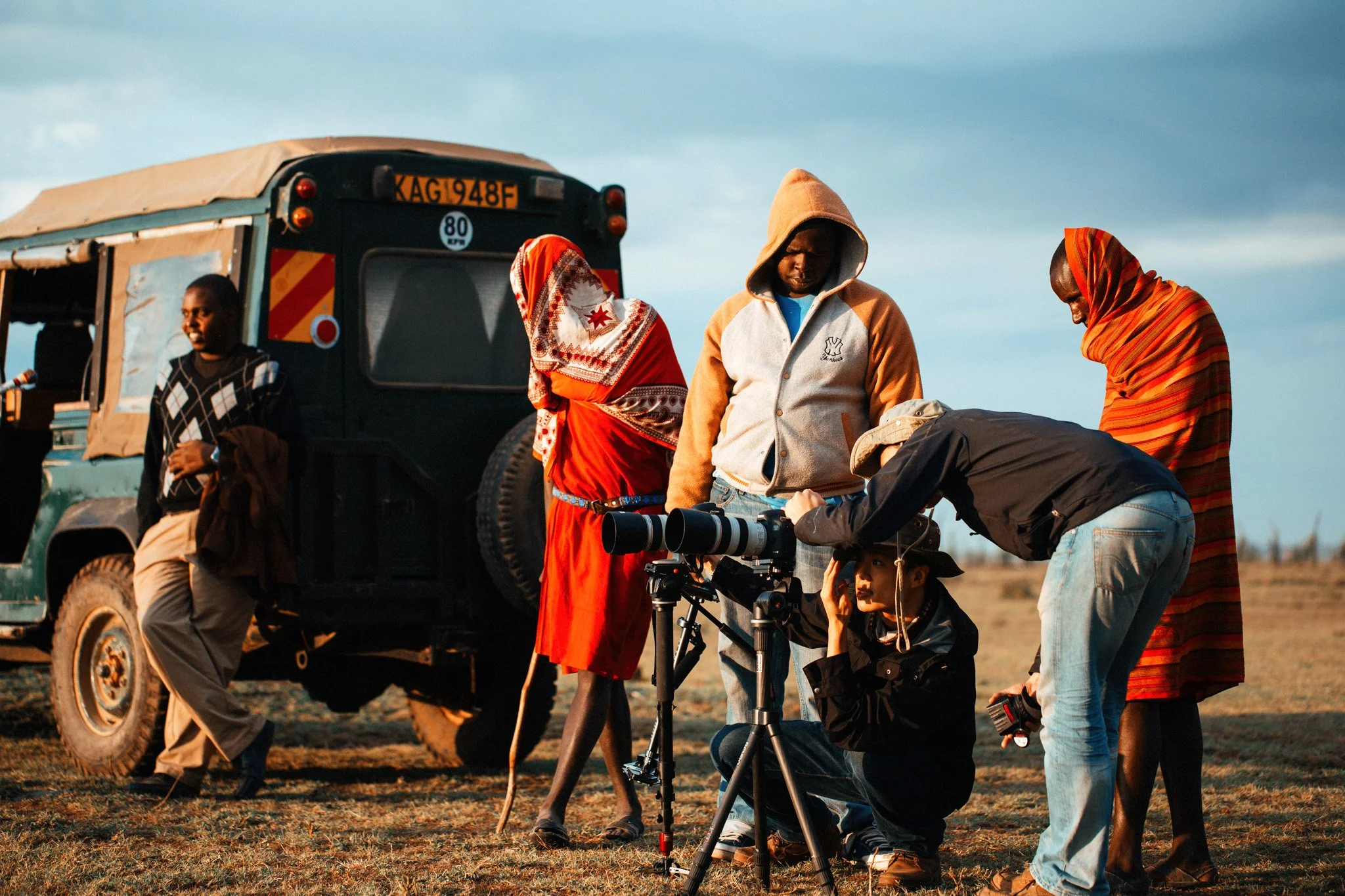 A group of people on a film or photography shoot outdoors, with a vehicle in the background. They are gathered around a camera drone on a tripod, and some are adjusting or working with equipment.