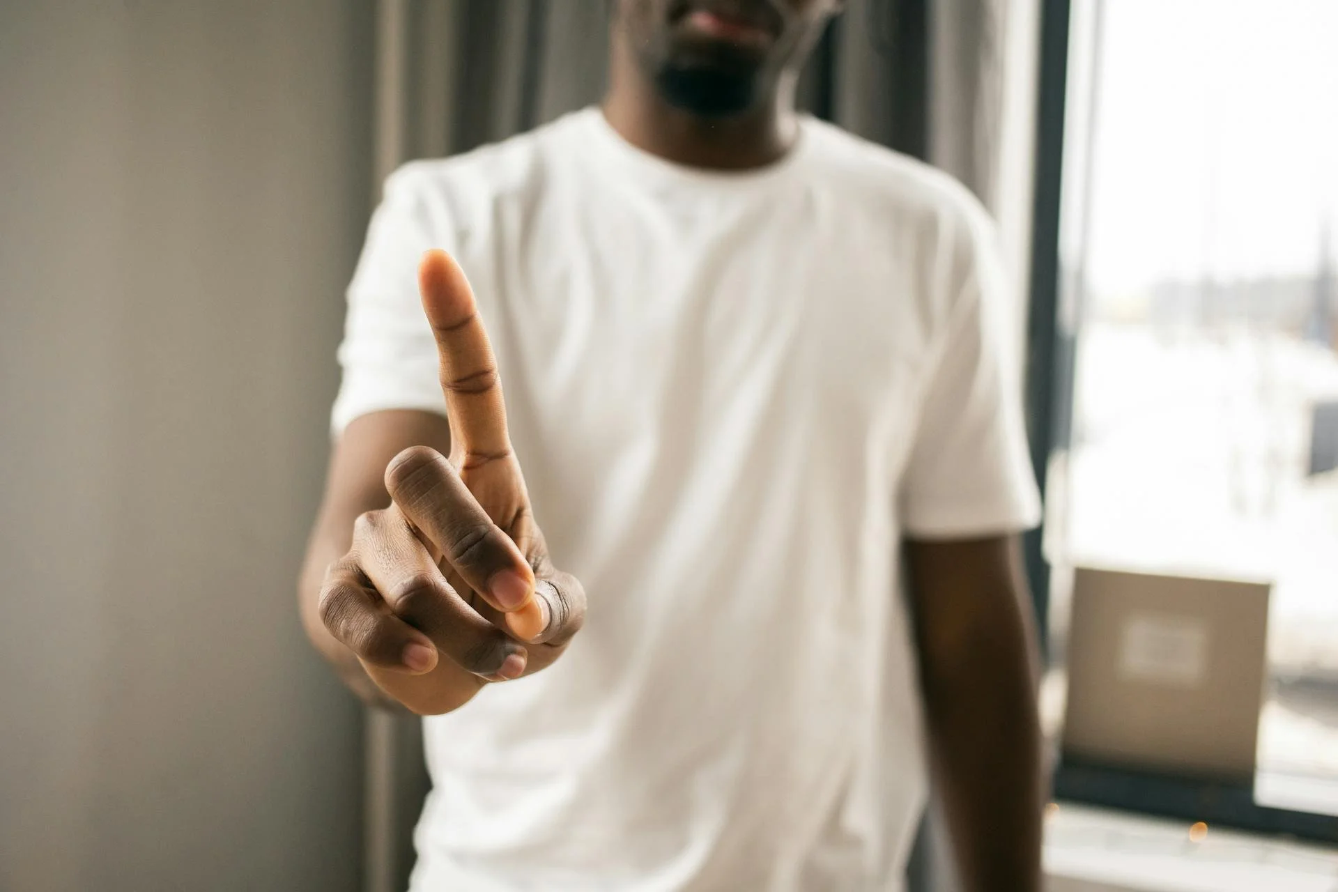 A person in a white t-shirt holding up their index finger in a pointing gesture. The focus is on the hand in the foreground, while the person's face is blurred in the background. The setting appears to be indoors with window light visible.