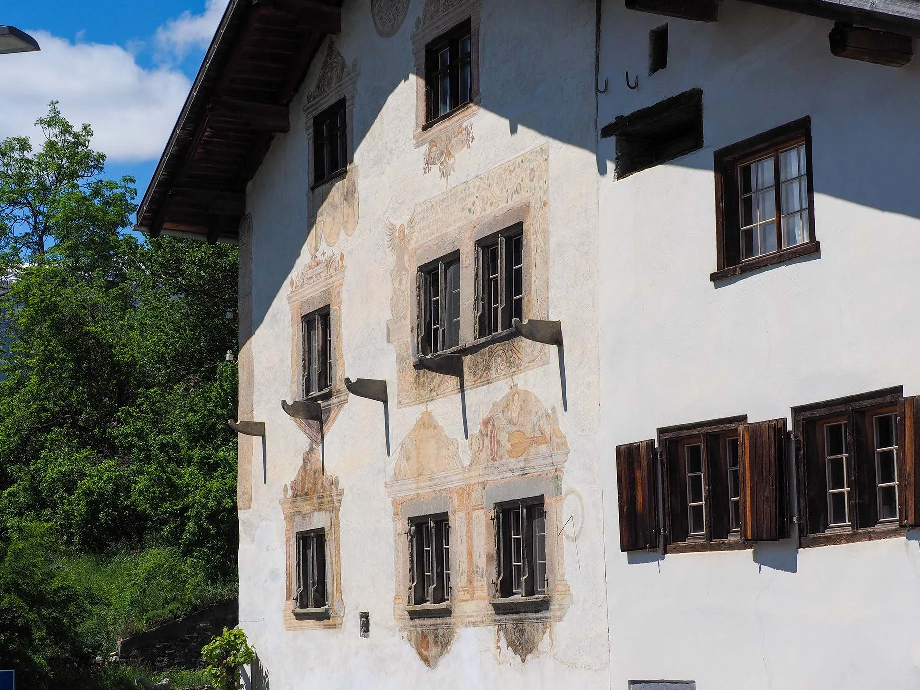 Ein altes, mehrstöckiges Haus mit weißen Wänden und dunklen Fensterrahmen, das teilweise mit Fresken verziert ist. Im Hintergrund grüner Baum und blauer Himmel mit Wolken.