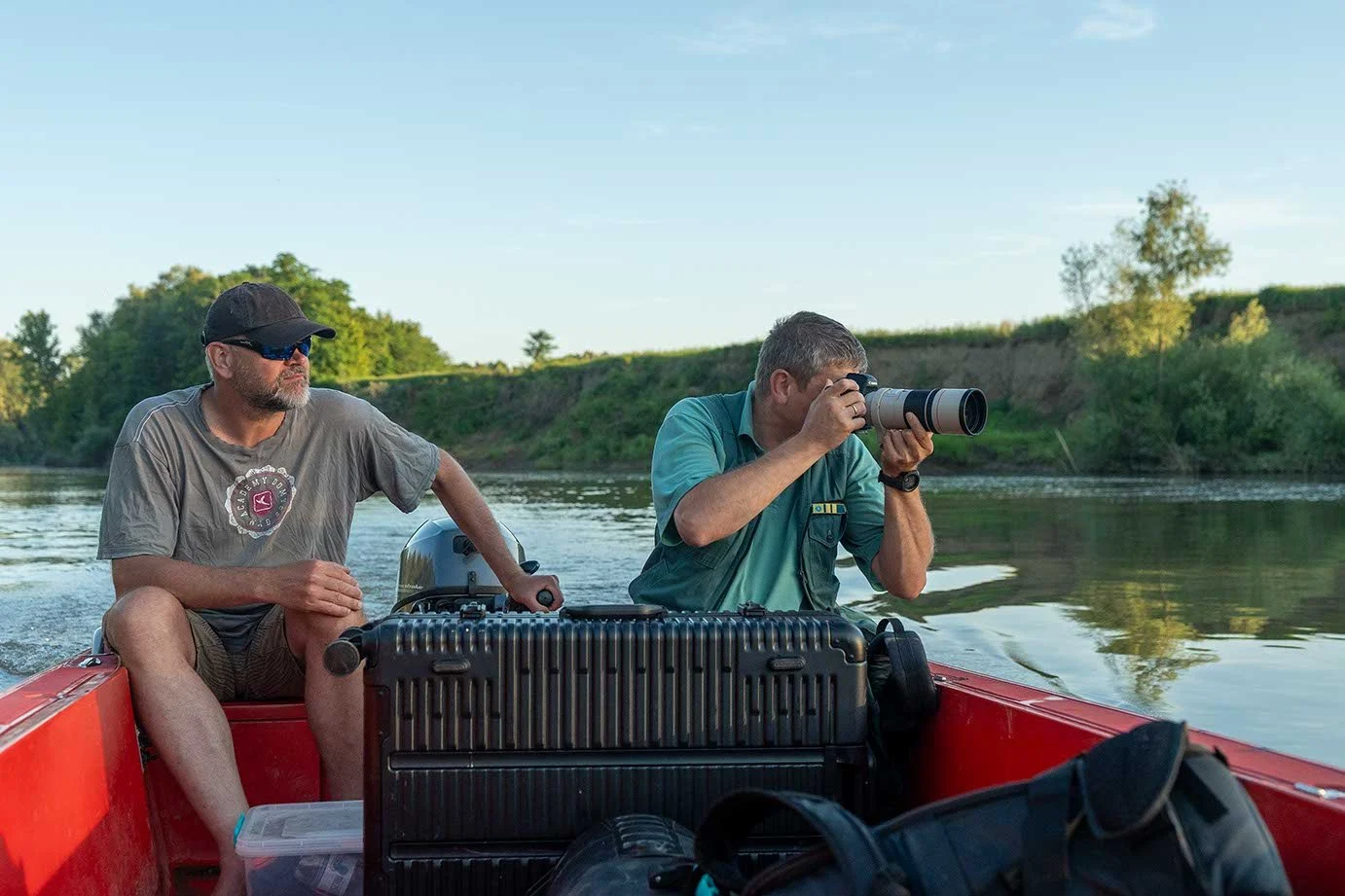 Zwei Männer auf einem roten Boot auf einem Fluss, einer schaut durch ein Fernglas, der andere sitzt hinter ihm mit einem Tanktop und Sonnenbrille, umgeben von Bootsgepäck, bei Sonnenuntergang oder -aufgang.