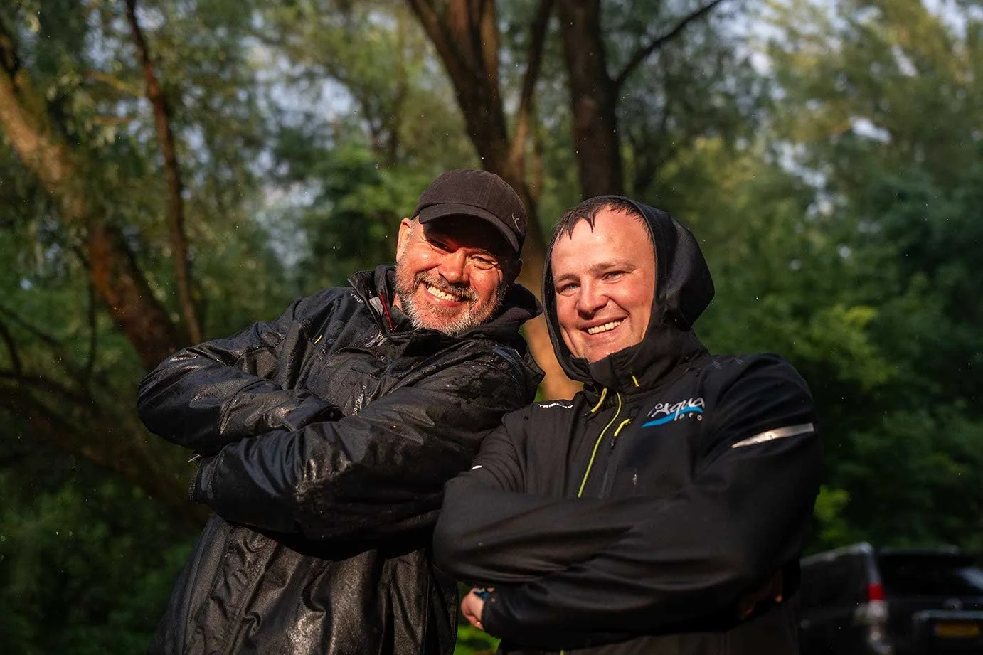 Zwei lachende Männer tragen wasserfestejacken im Grünen bei Regen, umarmt und freundlich.